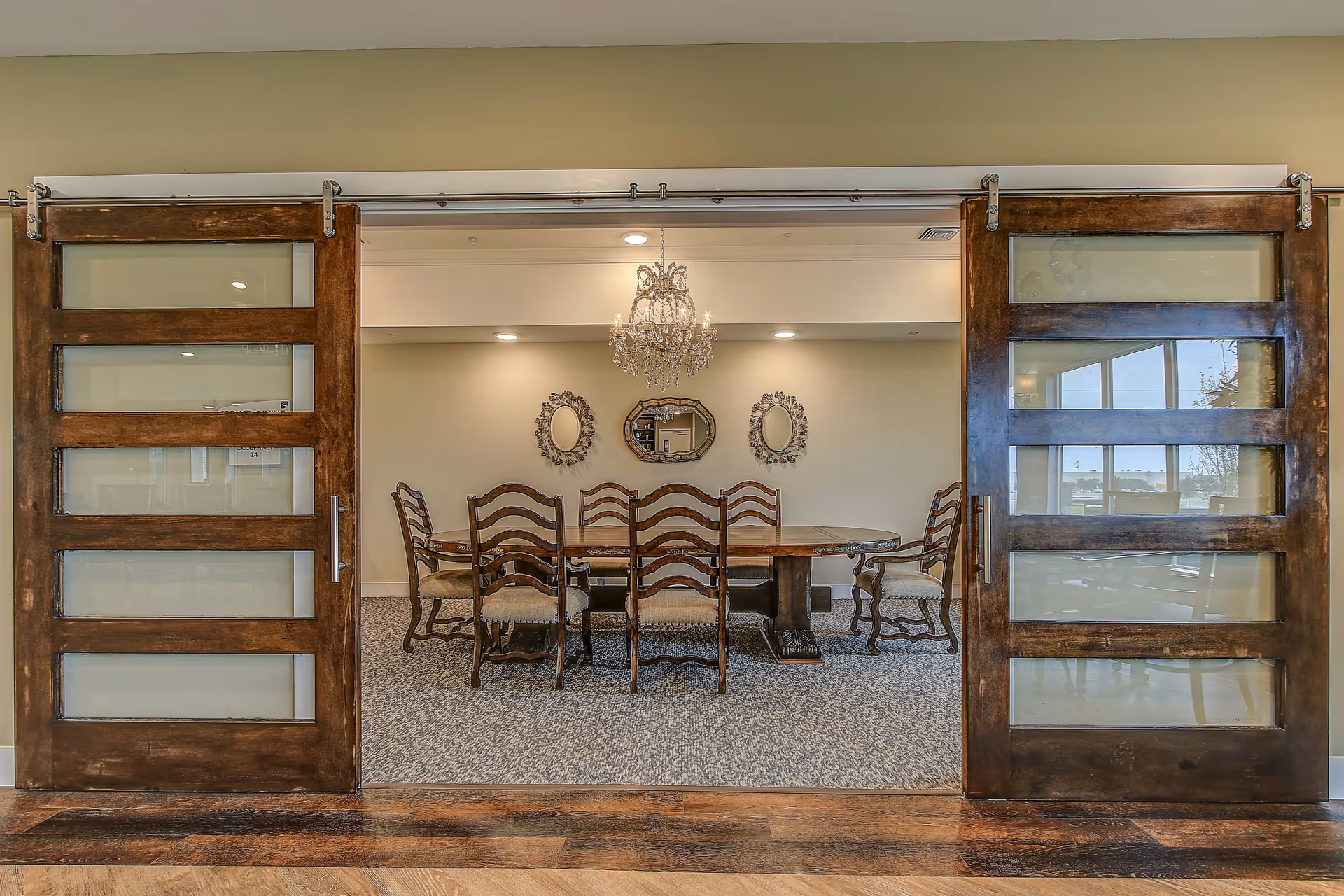 A dining room with a large wooden table surrounded by eight wooden chairs with upholstered seats. The room is viewed through two large sliding wooden doors with frosted glass panels. On the back wall, there are three decorative mirrors and a chandelier hanging from the ceiling. The floor is carpeted with a patterned design.