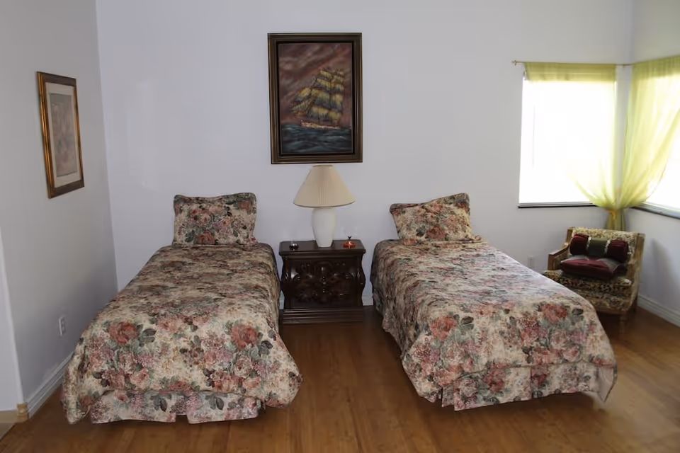 A bedroom with two twin beds covered in floral bedspreads. Between the beds is a wooden nightstand with a white lamp and small decorative items. On the wall above the nightstand is a framed painting of a sailing ship. To the right, there is a window with light green curtains and a cushioned armchair with pillows.