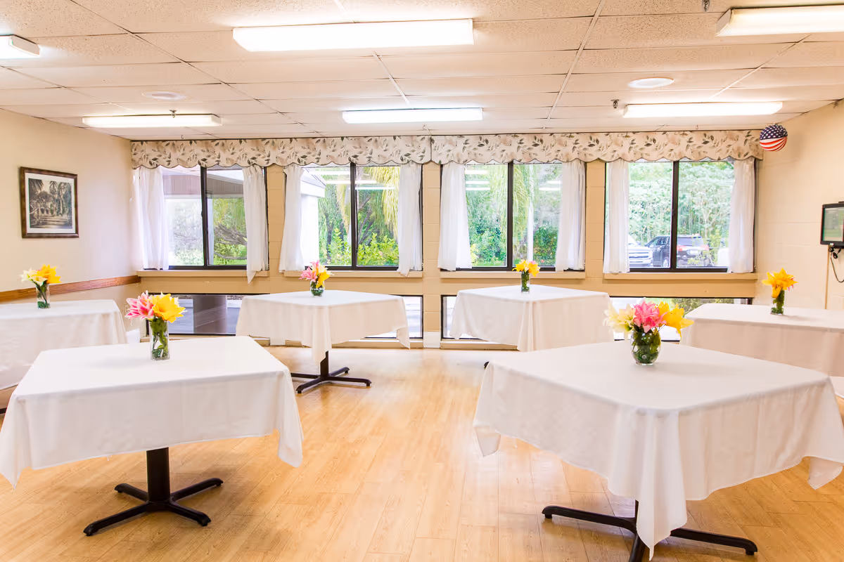 A bright dining room with six square tables covered with white tablecloths, each table decorated with a small vase of colorful flowers. Large windows with white curtains and floral valances let in natural light, and a framed picture hangs on the left wall. The floor is light wood, and fluorescent lights illuminate the ceiling.