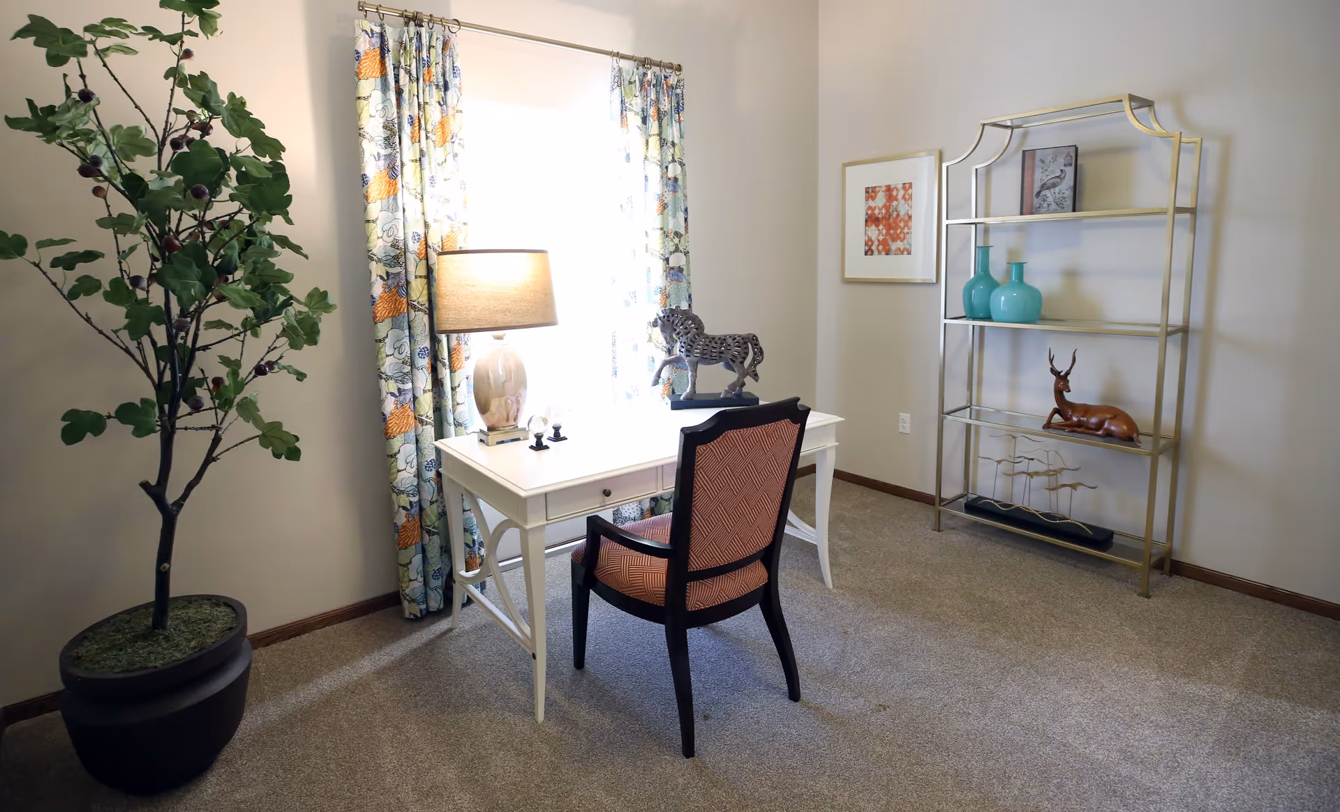 A small room with a white desk and a chair with a patterned cushion. On the desk is a lamp, a decorative horse sculpture, and some small items. A large potted plant is on the left side near a window with floral curtains. On the right side, there is a gold metal shelving unit with decorative vases and sculptures. The walls are light-colored and the floor is carpeted.