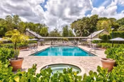 Outdoor swimming pool with an adjacent hot tub, lounge chairs and umbrellas, surrounded by landscaping and clubhouse buildings under a partly cloudy sky.