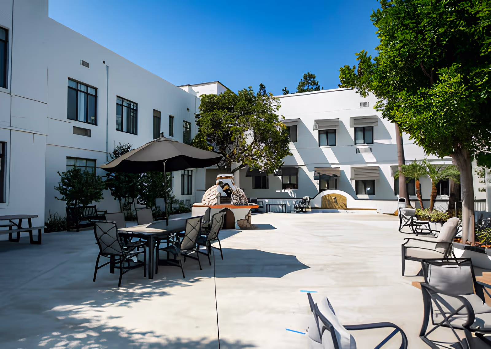 Outdoor courtyard area of French Park Care Center with patio tables and chairs, a large umbrella, trees providing shade, and a white building with multiple windows in the background under a clear blue sky.