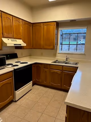 A kitchen with wooden cabinets, a white electric stove with four burners, a microwave, a double sink under a window with blinds, and beige tiled flooring.