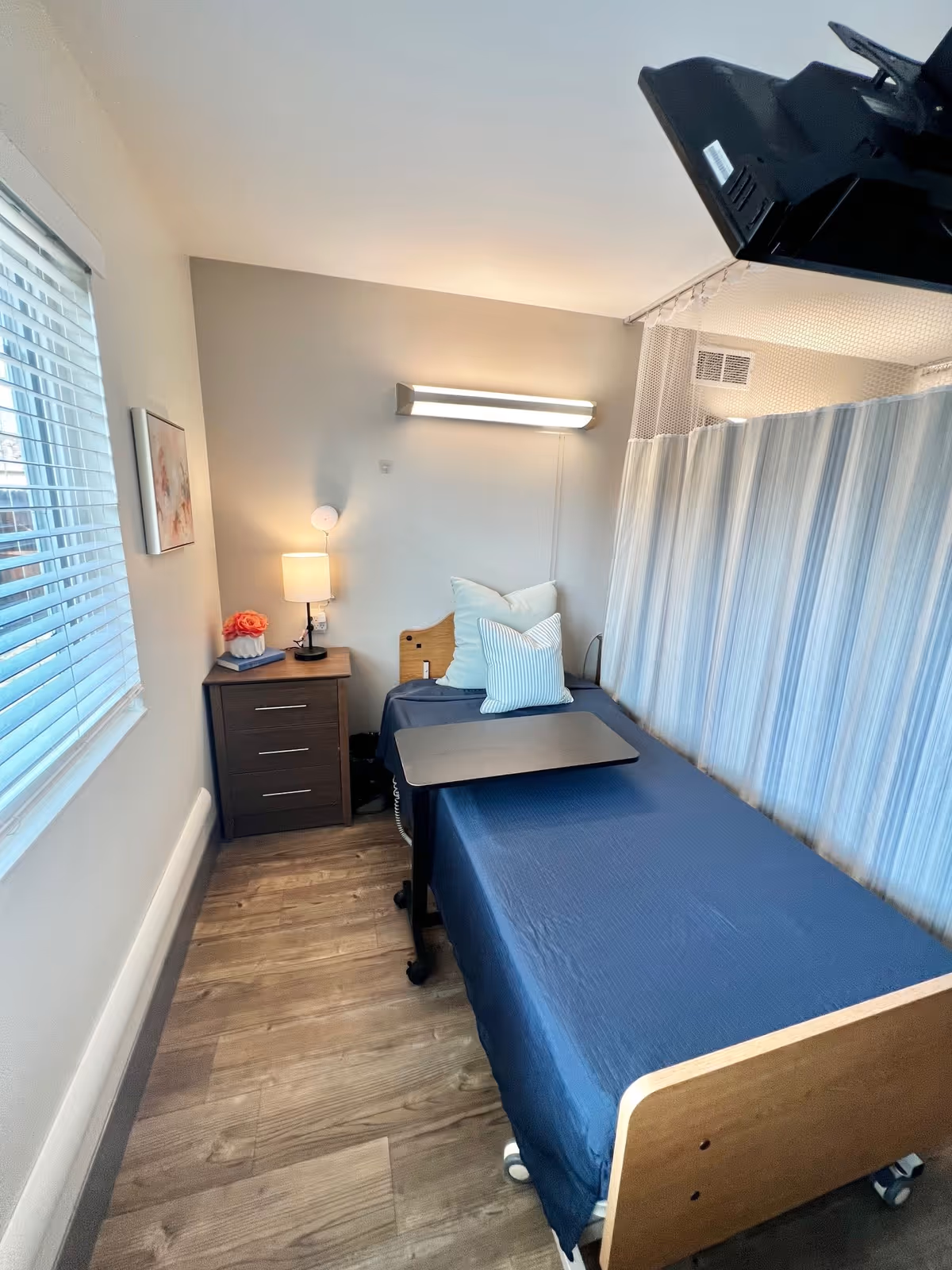 A small healthcare facility room with a single bed covered in a dark blue sheet, two pillows, a bedside table with a lamp and a small flower arrangement, a window with blinds, and a privacy curtain on one side.