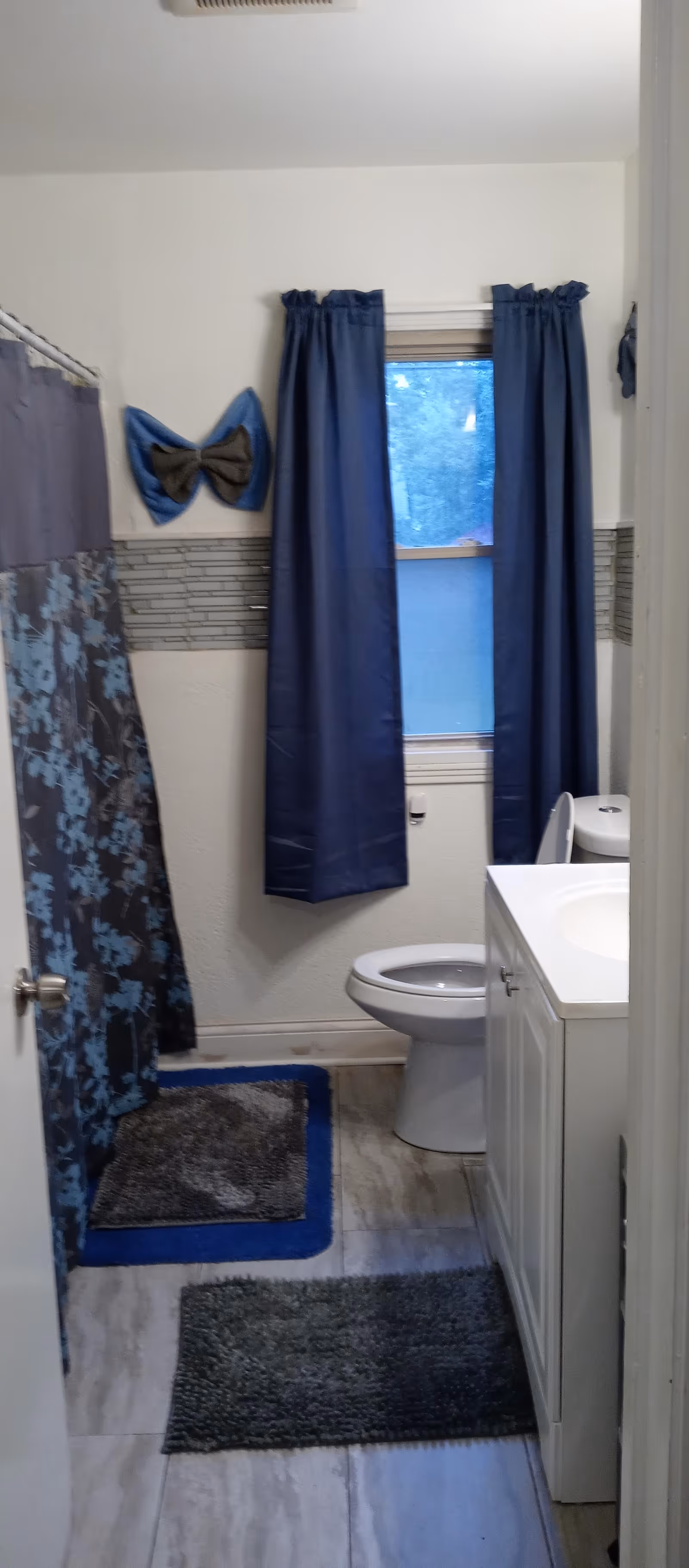 A small bathroom with a white toilet and white vanity with sink on the right side. There is a window with blue curtains above the toilet. On the left side, there is a shower with a floral patterned shower curtain in shades of blue and gray. The floor has gray tiles with two dark gray and blue bath mats. A decorative blue and gray bow hangs on the wall near the window.