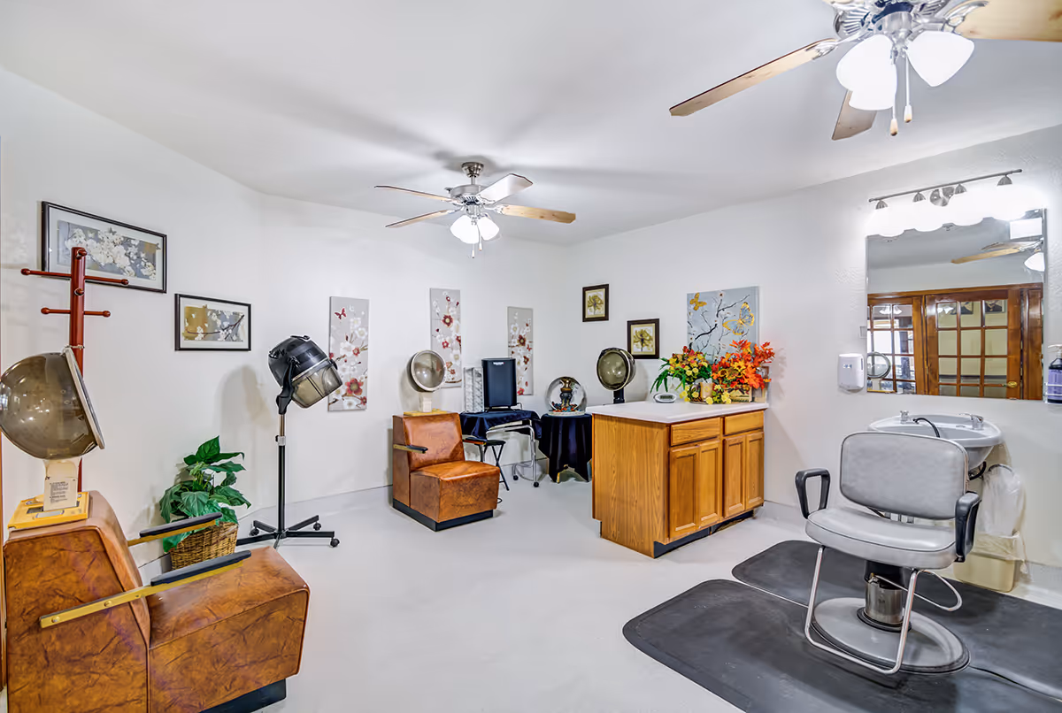 Interior of a salon room in a senior living facility with vintage hair drying chairs, hair dryers, a salon chair in front of a mirror with a sink, wooden cabinets, decorative wall art, and ceiling fans with lights.
