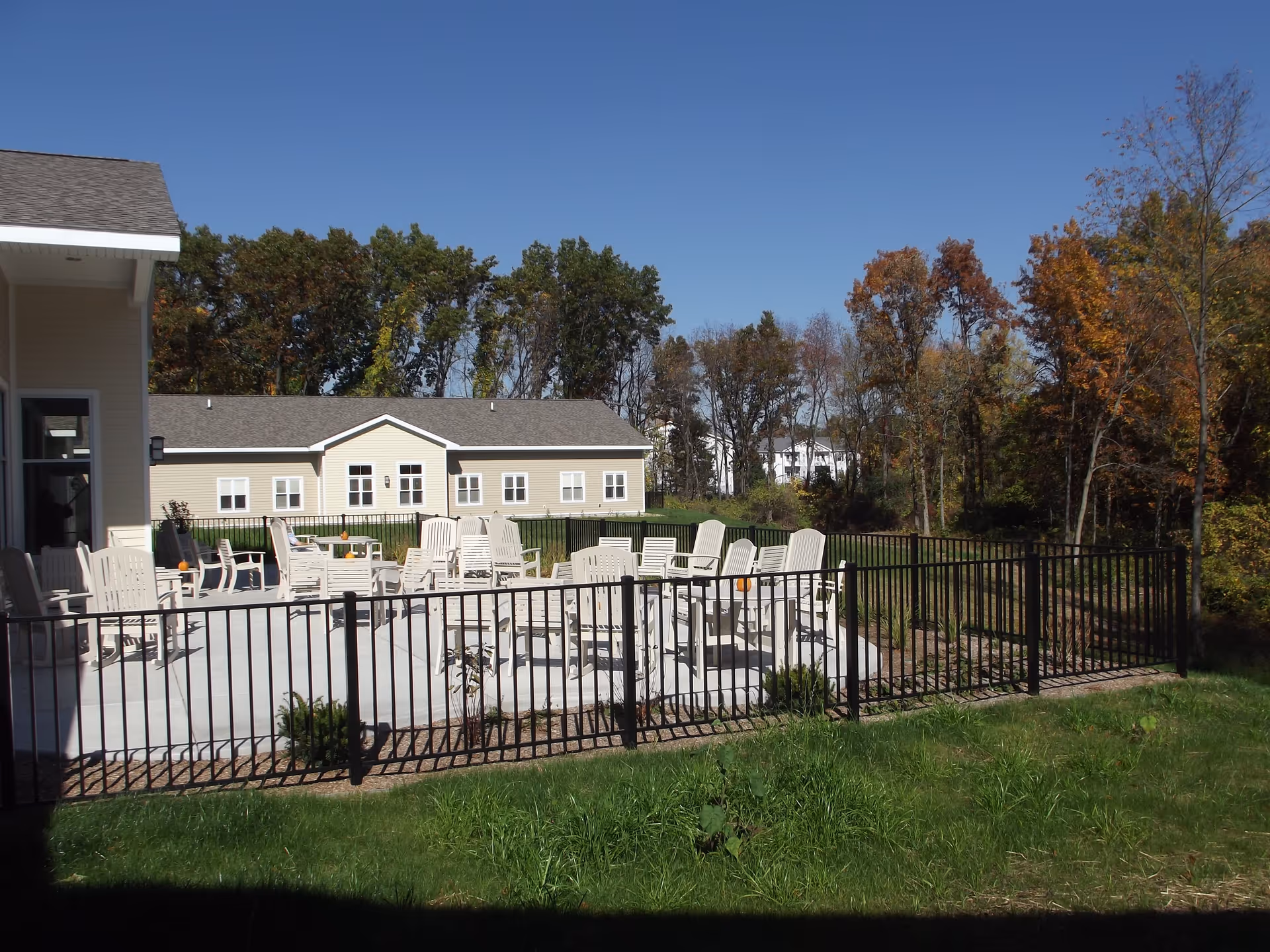 Fenced outdoor patio with multiple white chairs and tables in front of a single-story building and trees.