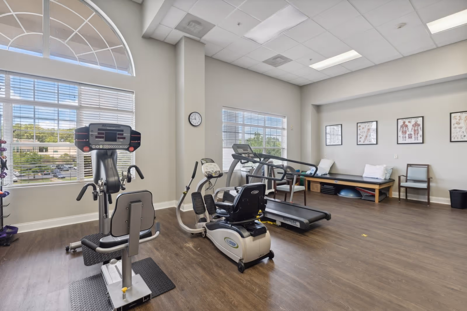 A bright exercise room with large windows and wood flooring, featuring various fitness equipment including a recumbent bike, treadmill, and a multi-exercise machine. There is a bench with pillows and anatomical charts on the wall, along with a clock and chairs.
