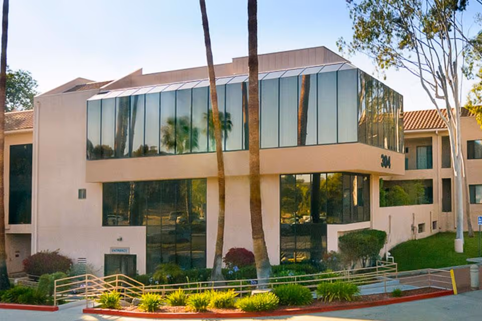 Exterior front of a multi-story senior living facility with large reflective glass windows, palm trees, and a landscaped entrance ramp.