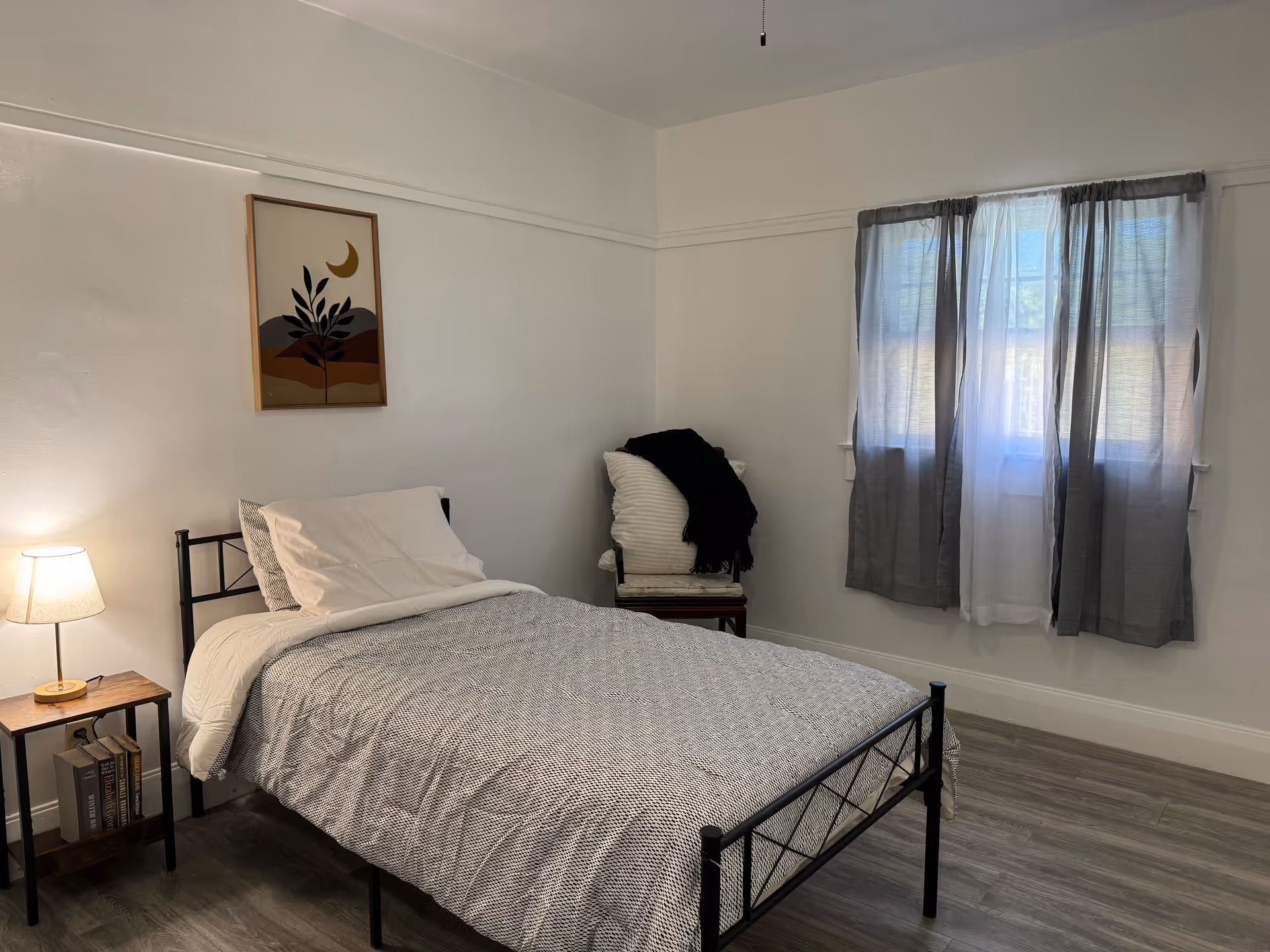A simple bedroom with a single bed featuring a gray and white patterned bedspread and white pillows. A small wooden side table with a lamp and books is next to the bed. A chair with a white cushion and a black blanket is placed near a window with gray and white curtains. A framed abstract art piece hangs on the white wall above the bed. The floor is wood with a light finish.