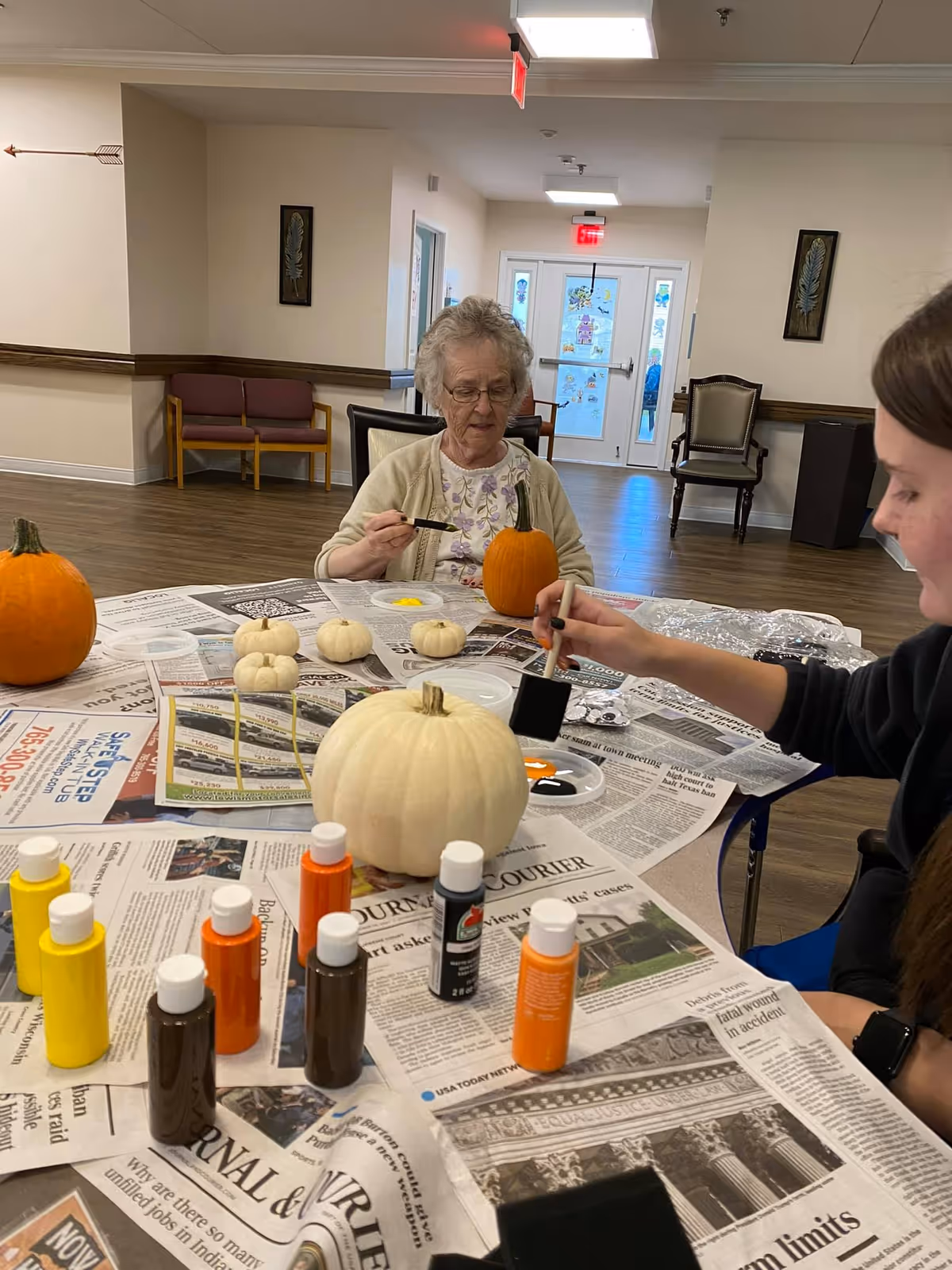 An elderly woman and a younger woman sitting at a table covered with newspapers, painting small pumpkins with brushes and various colors of paint bottles in a well-lit room with wooden flooring and chairs in the background.