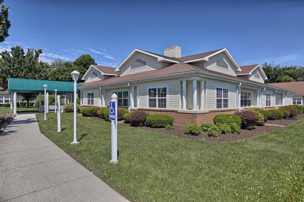 Exterior view of a single-story senior living facility building with beige siding and brick accents, surrounded by green grass and bushes under a clear blue sky. A concrete walkway leads to a green canopy entrance with white lamp posts along the path. A handicap parking sign is visible near the walkway.