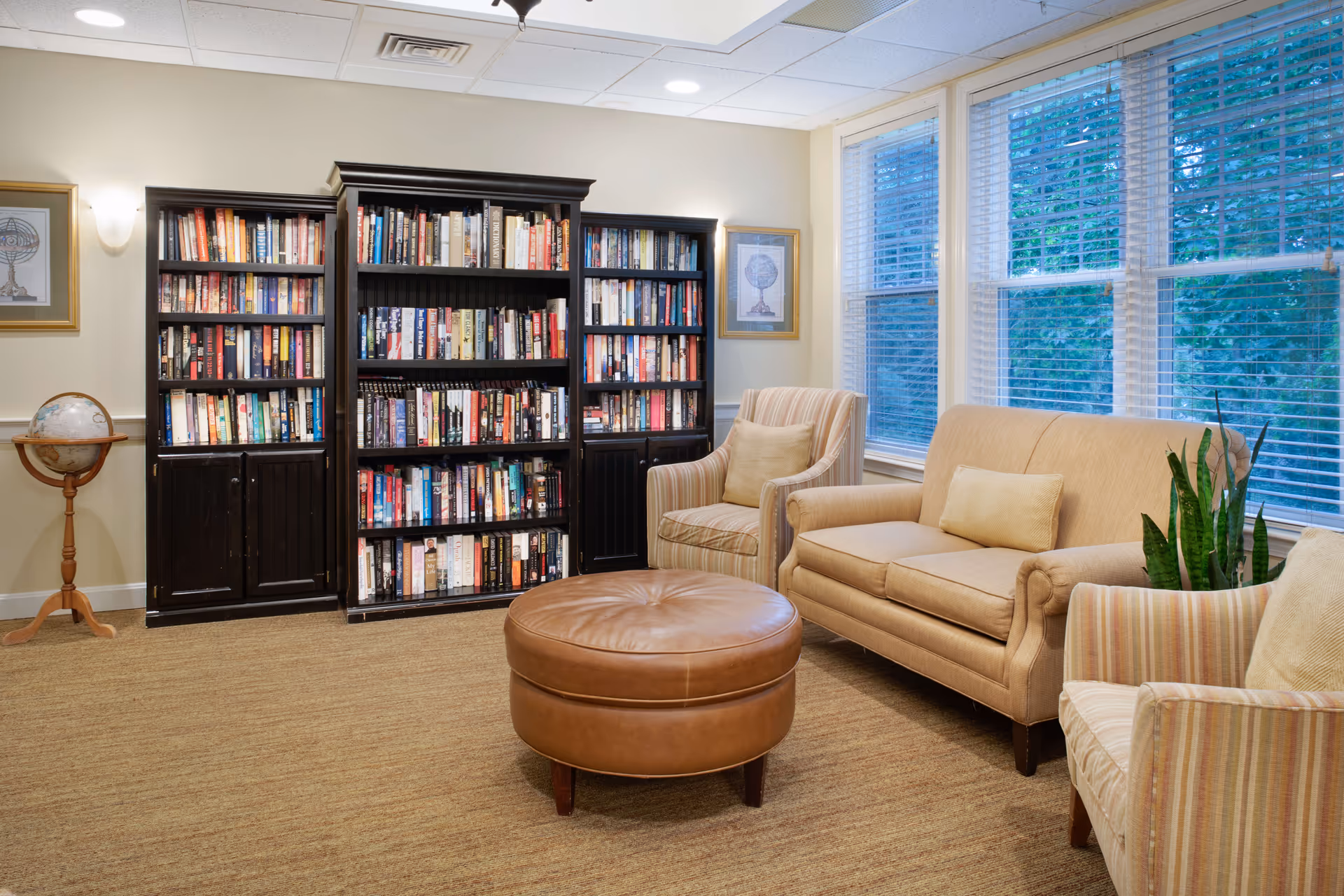 Cozy lounge with bookshelves, armchairs, a sofa, and a round leather ottoman in front of large windows.