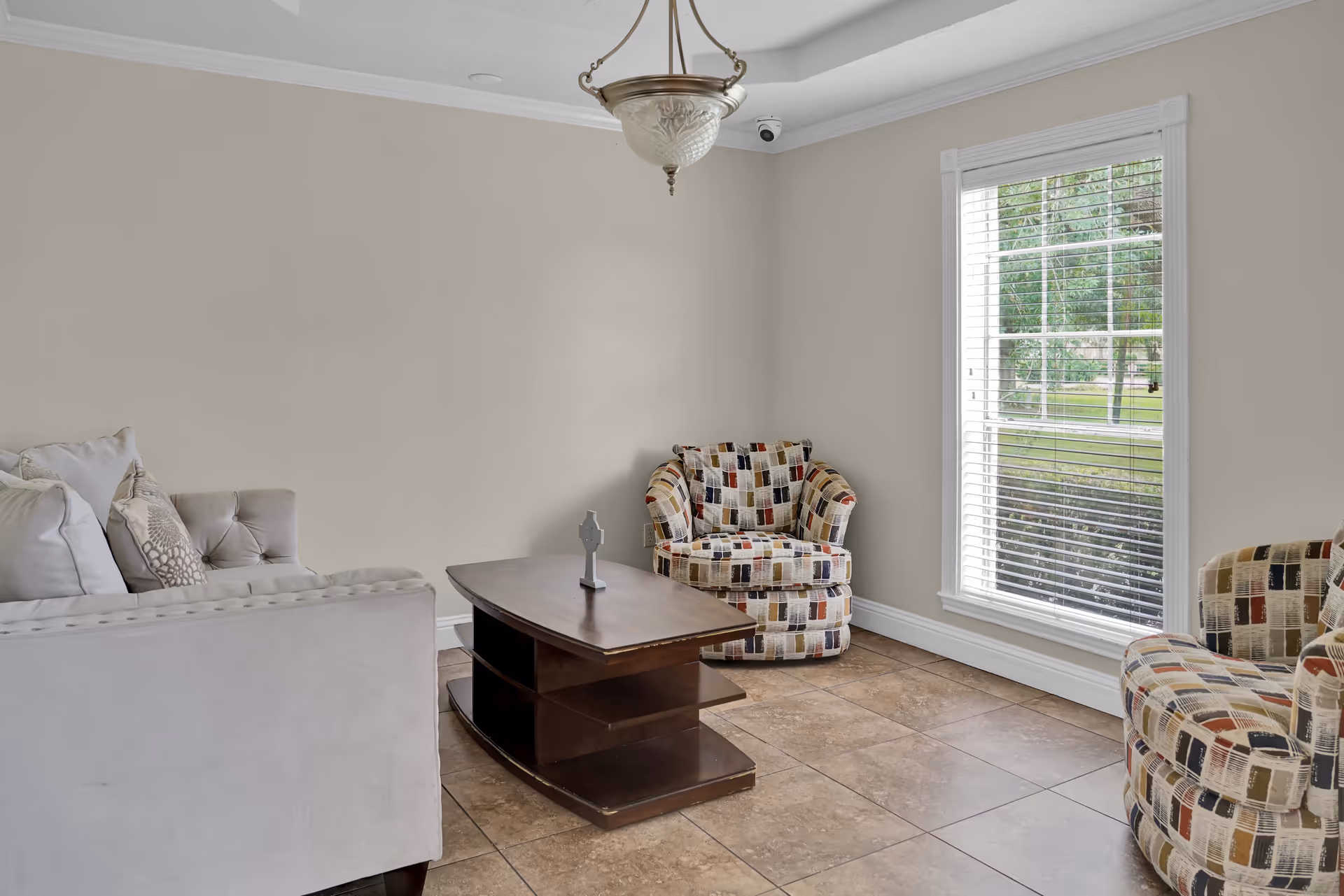A cozy living room with a beige sofa adorned with pillows, two patterned armchairs, a dark wooden coffee table with a decorative item on top, a large window with white blinds letting in natural light, beige walls, and a ceiling light fixture.
