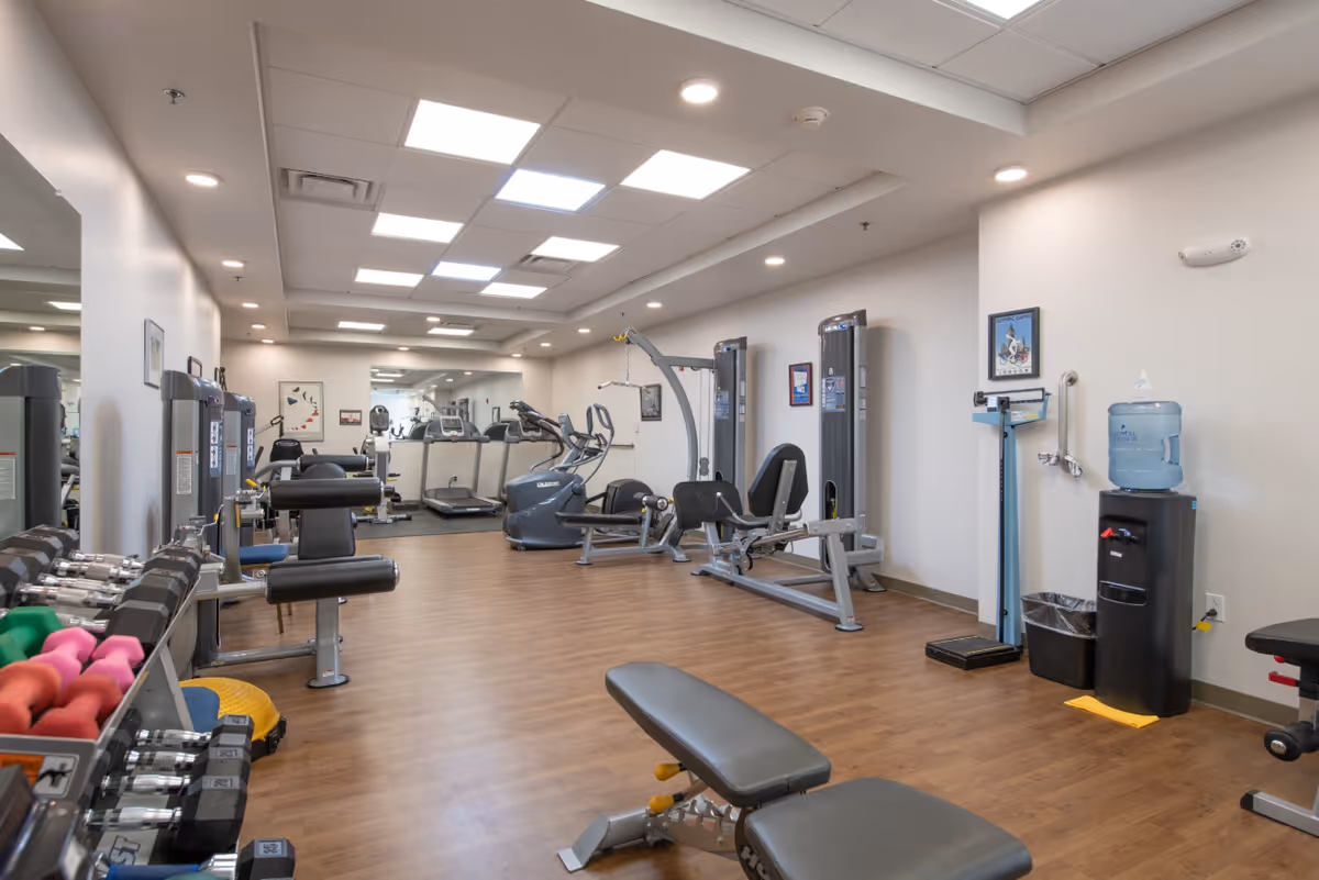 A well-lit fitness room with various exercise equipment including dumbbells, weight machines, treadmills, and stationary bikes. The room has wooden flooring, white walls with framed pictures, and a water dispenser with a scale nearby.