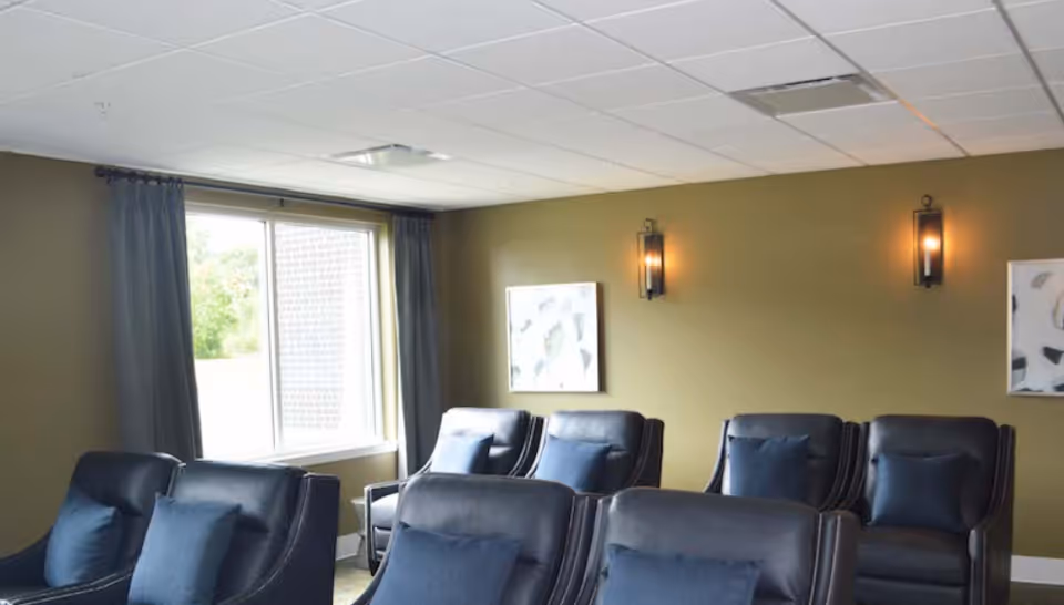 A communal lounge with several dark leather recliners, an olive-green wall with sconces and artwork, and a window with curtains.