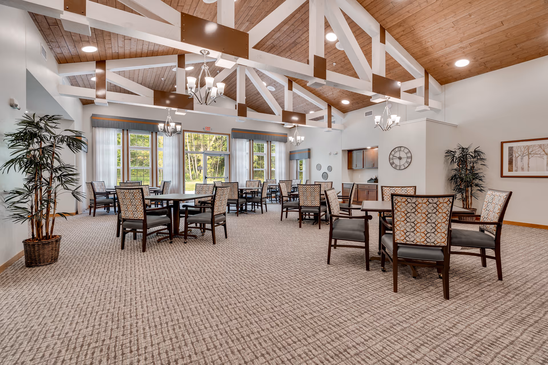 A spacious senior living dining room with multiple tables and chairs arranged neatly on a patterned carpet. The room features high vaulted ceilings with exposed wooden beams and chandeliers. Large windows allow natural light to brighten the space, and there are potted plants and framed artwork on the walls.