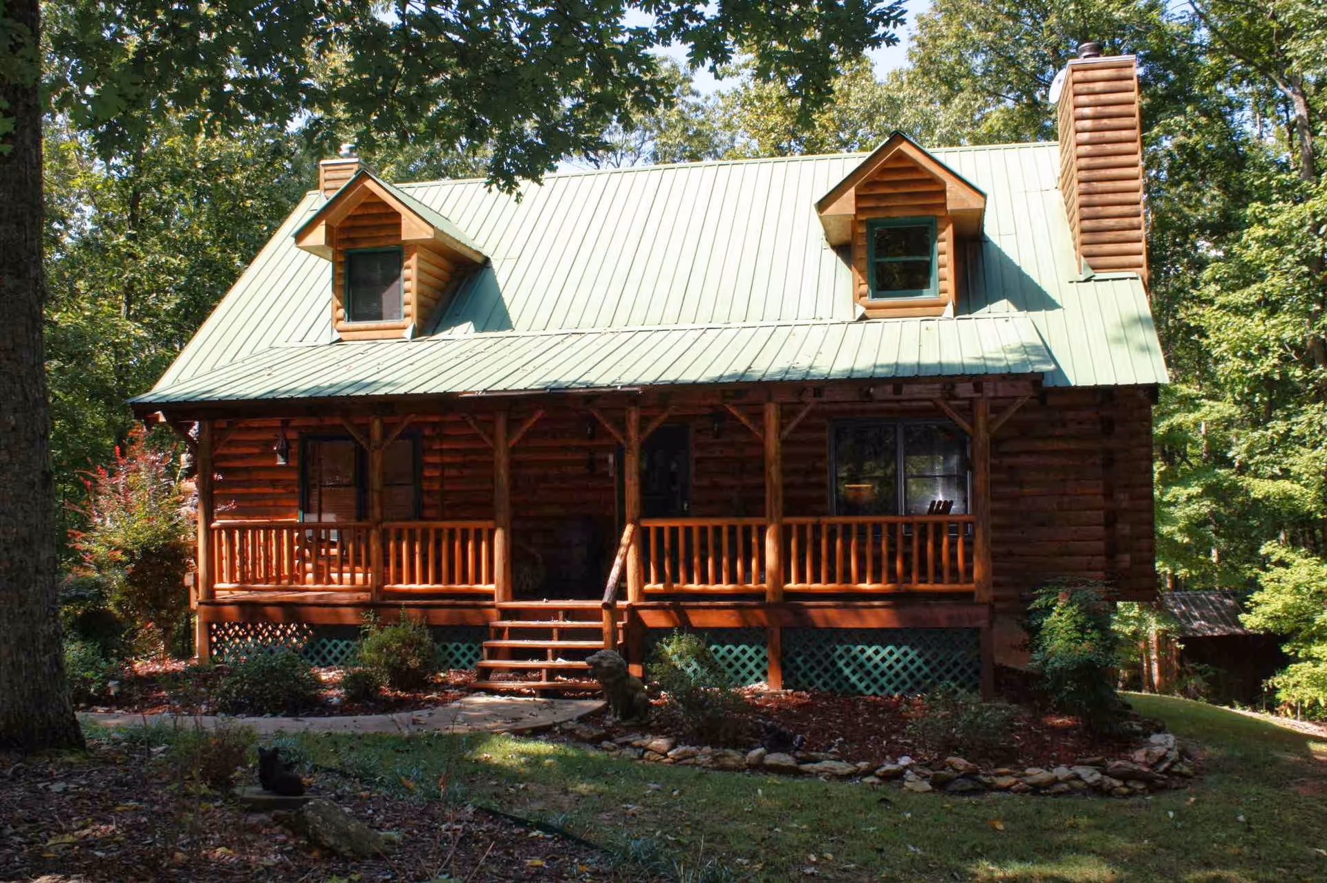Front exterior view of a rustic log cabin with a green metal roof, two dormer windows, a chimney, and a wooden porch surrounded by trees and landscaping.
