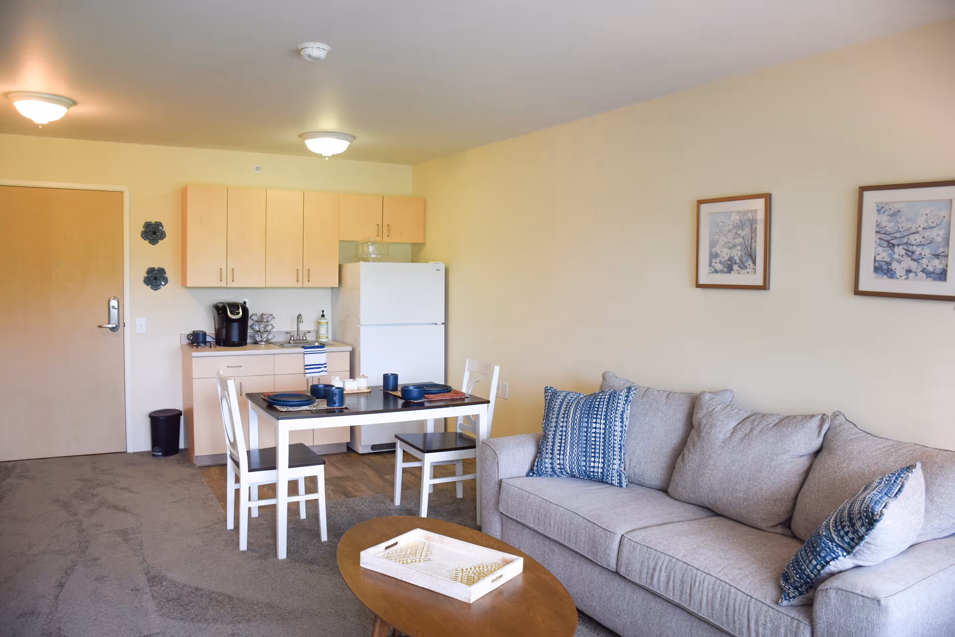 A cozy living area with a light gray sofa adorned with blue and white patterned pillows. In front of the sofa is a wooden coffee table with a chessboard on it. To the left, there is a small dining table set with plates and cups, accompanied by two white chairs. Behind the dining area is a compact kitchen with light wood cabinets, a white refrigerator, a coffee maker, and a sink. The walls are light-colored with two framed floral artworks hanging above the sofa. The room has carpeted flooring and ceiling lights.