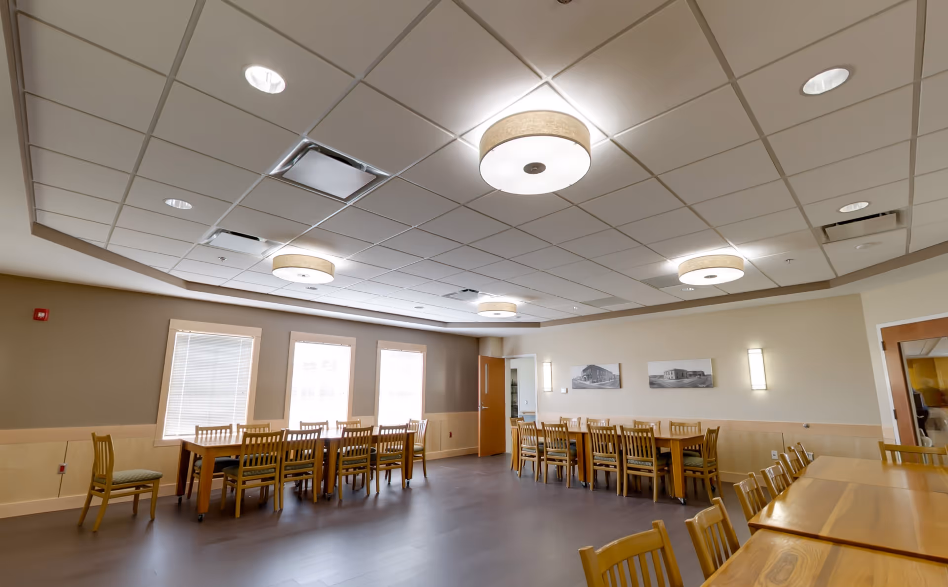 Bright communal dining room with multiple wooden tables and chairs under recessed ceiling lights.