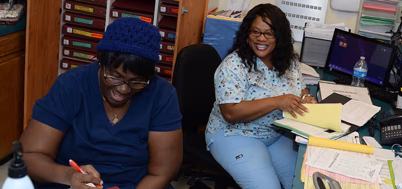 Two women in a medical office setting; one is wearing navy blue scrubs and a blue knit cap, writing with a red pen, while the other is wearing light blue scrubs with a pattern, smiling and organizing papers at a cluttered desk with a computer and various documents.
