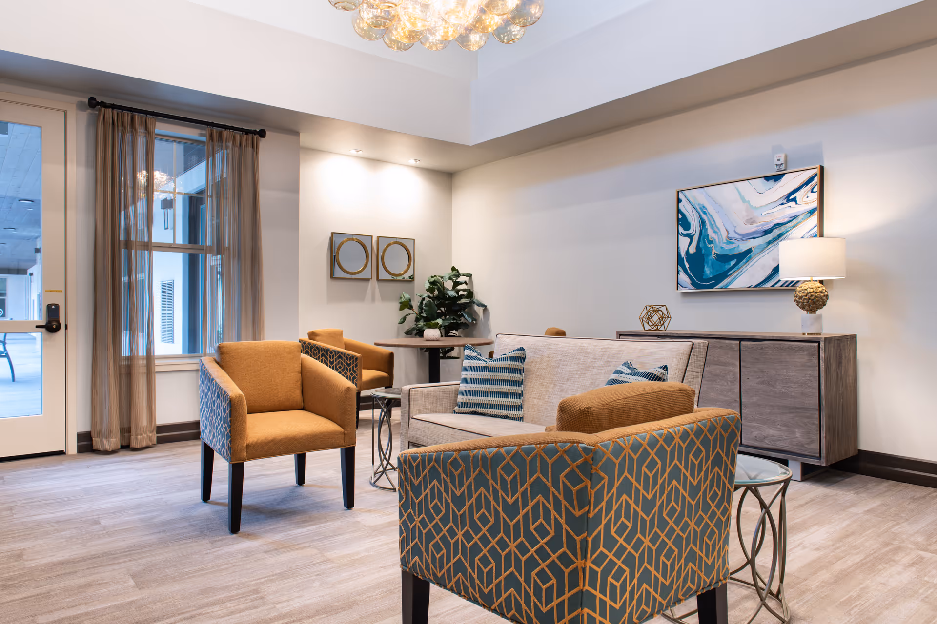 Bright seating area in a senior living lounge featuring patterned armchairs, a sofa, side tables, artwork, and a lamp.