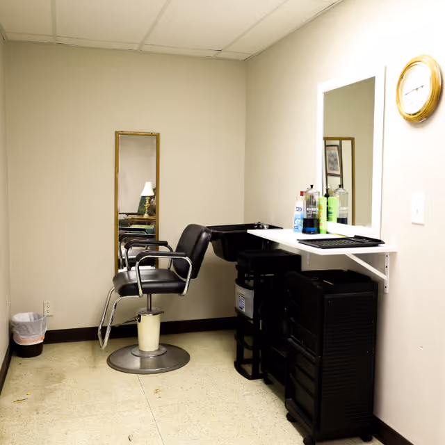 A small hair salon area inside a room with a black salon chair, a wall-mounted mirror, a tall narrow mirror on the adjacent wall, a black sink for washing hair, and various hair care products on a white shelf. There is a clock on the wall and a small trash can in the corner.