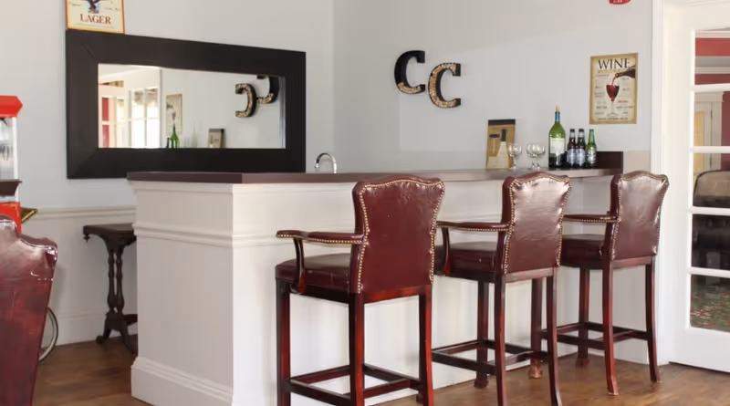 Interior view of a bar area with three high-backed leather chairs in front of a white counter with a dark wooden top. Behind the counter are bottles of wine and beer, wine glasses, and wall decorations including a large rectangular mirror, two letter 'C' wall hangings, and a framed wine poster. The floor is wooden and there is a glass door to the right.