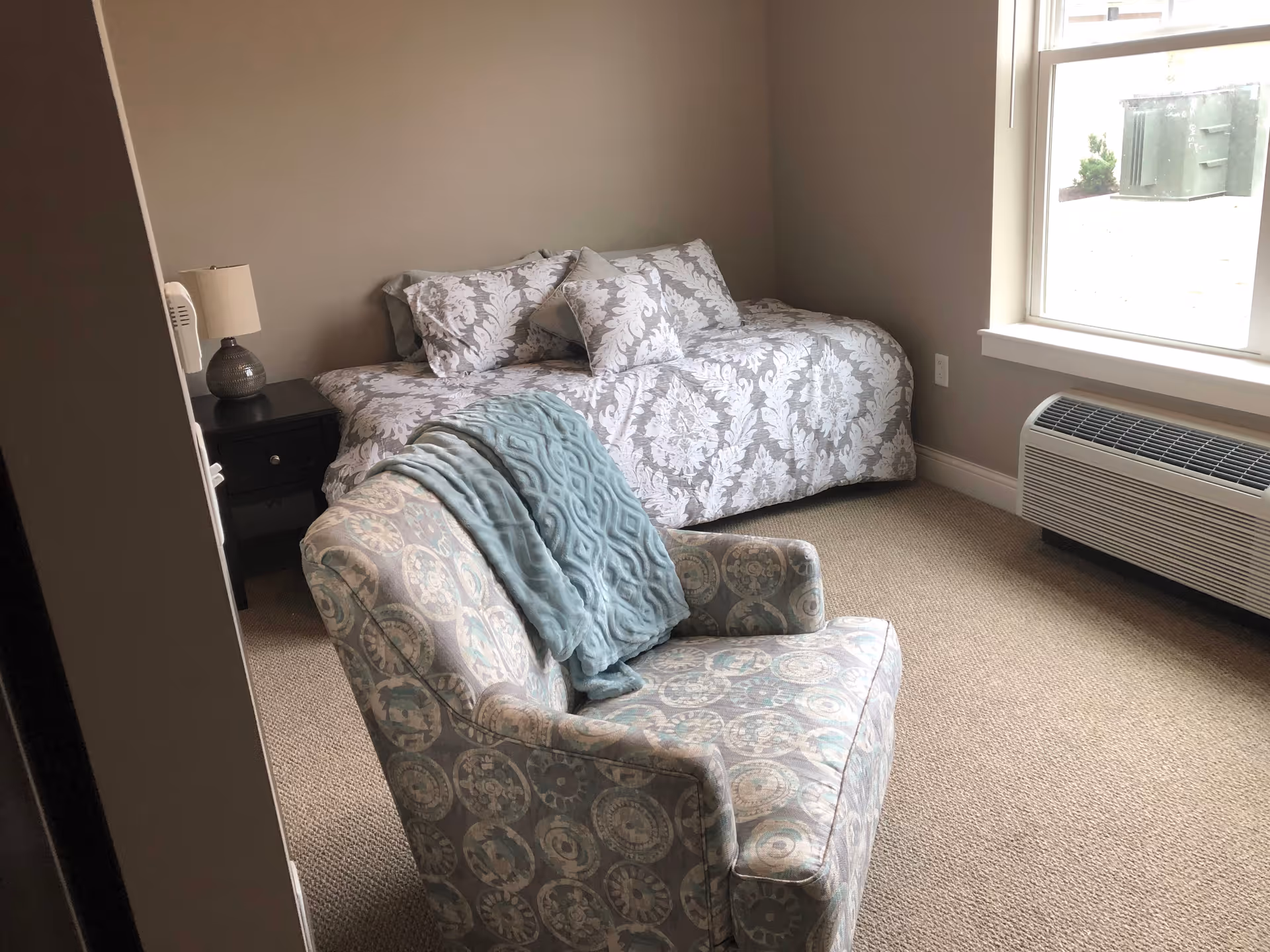 A small bedroom with a single bed covered in a patterned white and gray comforter and pillows. Next to the bed is a black nightstand with a lamp. In the foreground, there is a patterned armchair with a blue throw blanket draped over it. A window on the right side lets in natural light, and below the window is a heating/cooling unit.