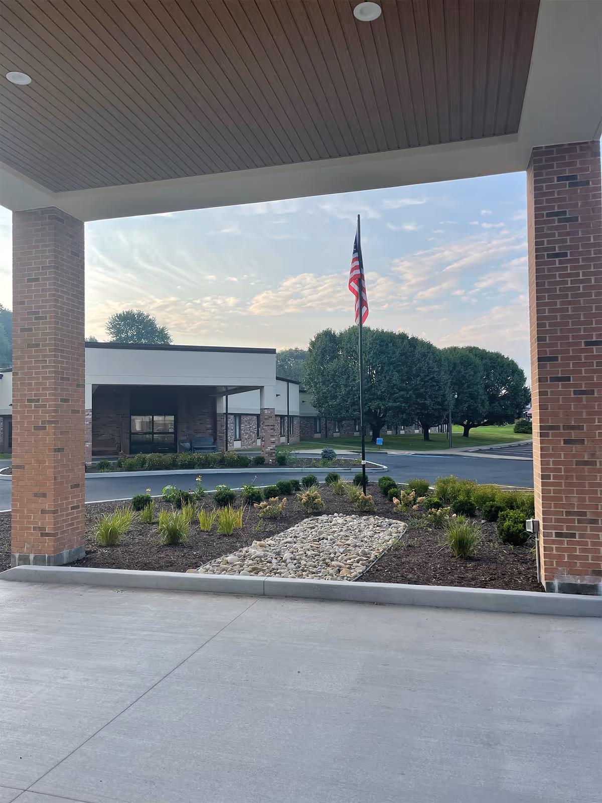 View from under a covered entrance area of a healthcare facility showing a landscaped garden with rocks and plants, an American flag on a flagpole, a driveway, and the building with brick pillars and trees in the background under a partly cloudy sky.