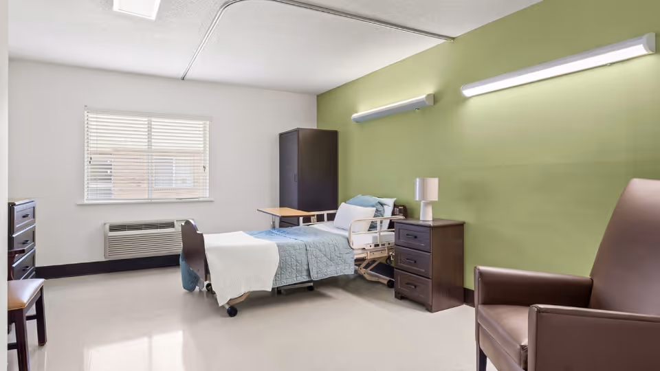 A simple, clean patient room with a single hospital bed covered with a blue quilt and white blanket. The room has a green accent wall with two horizontal lights, a wooden nightstand with a lamp, a dark wooden wardrobe, a brown leather armchair, a wooden chair, and a window with blinds letting in natural light.