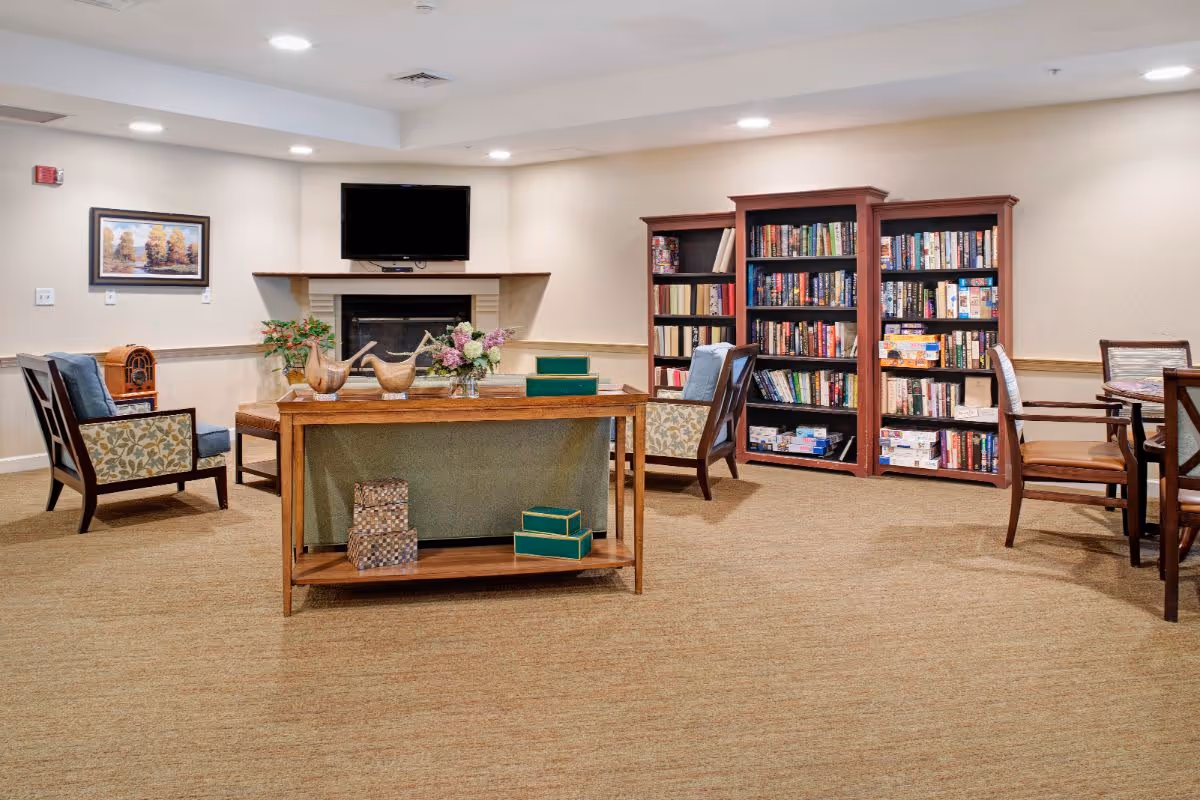 A cozy living room area in a senior living facility featuring a fireplace with a TV mounted above it, two wooden bookshelves filled with books and board games, a wooden table with decorative items and flowers, and several upholstered chairs arranged around the room.