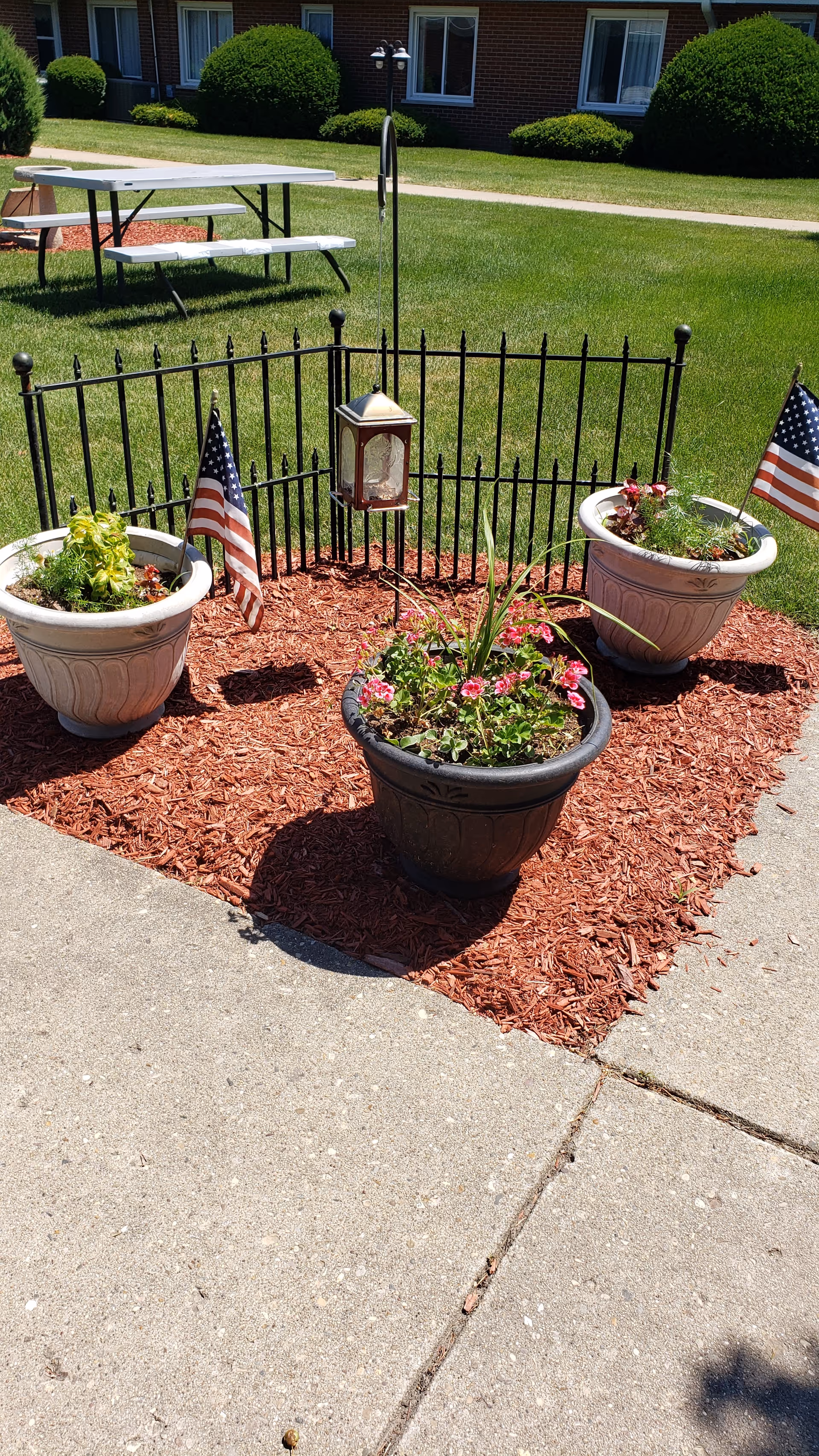 Outdoor garden area with three large flower pots containing various plants and flowers, two small American flags placed in the pots on the left and right. A small black metal fence encloses a lantern hanging from a hook in the center. In the background, there is a grassy lawn with trimmed bushes and a white picnic table with benches near a brick building with windows.