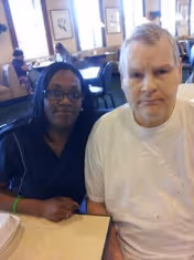 Two adults seated at a table in a communal dining area with other diners and windows in the background.