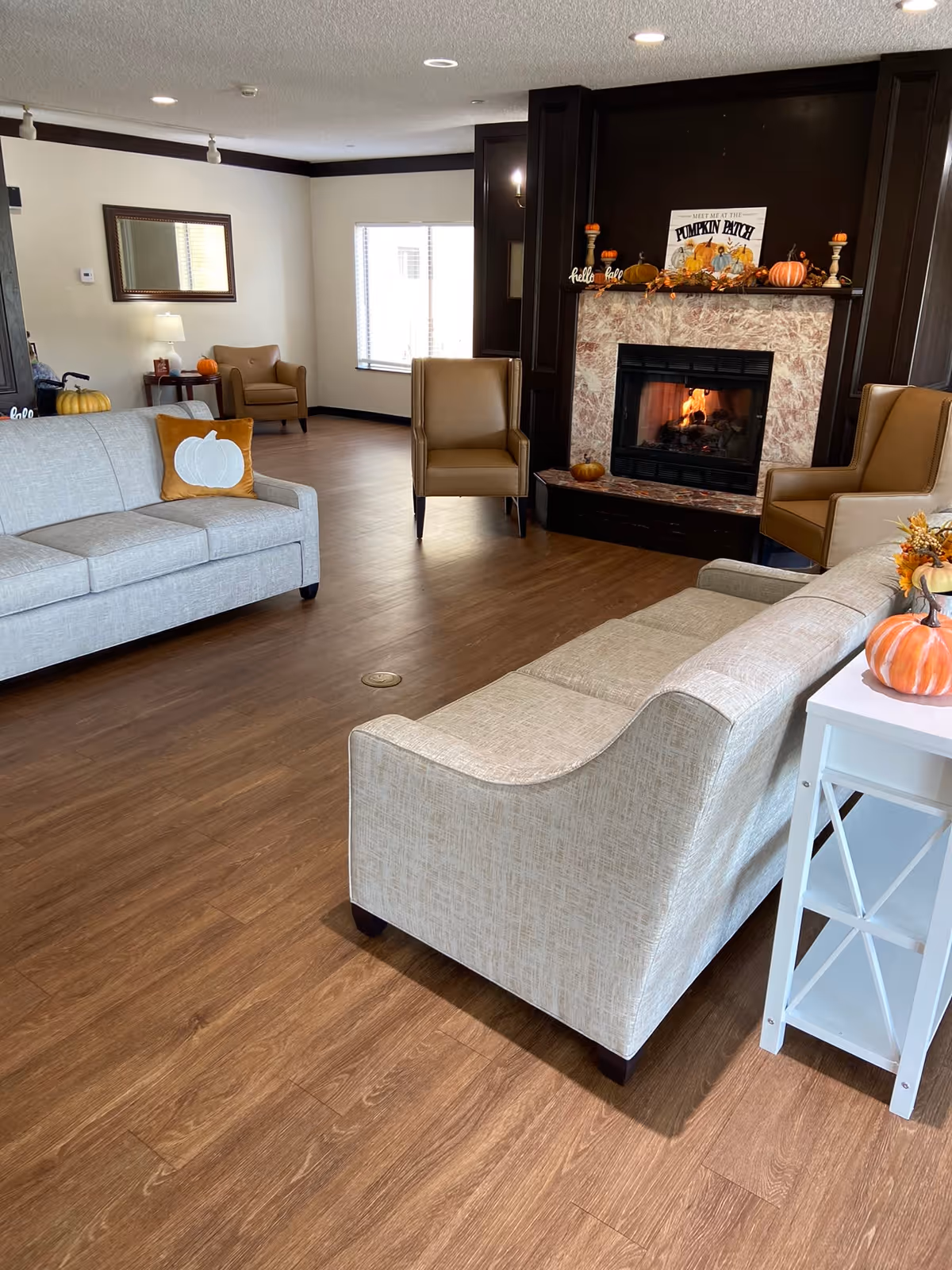A cozy senior living room with two beige sofas, two brown armchairs, a fireplace decorated with pumpkins and fall-themed items, and a wooden floor. The room has a window letting in natural light and a white side table with a pumpkin decoration.