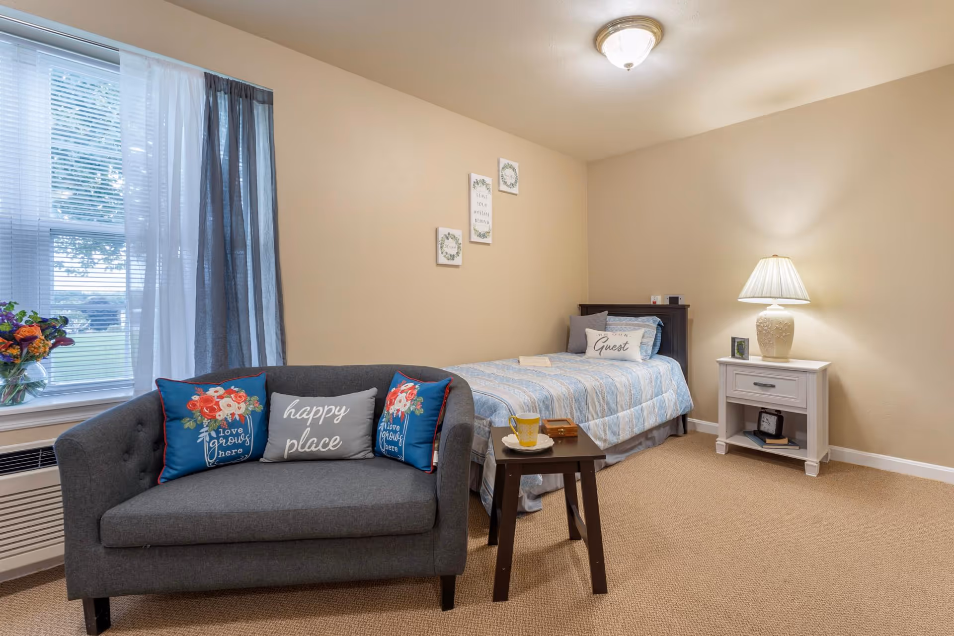 A cozy bedroom with a single bed covered in a blue and white striped quilt, a gray loveseat with decorative pillows, a small wooden side table with a cup and saucer, a white nightstand with a lamp, and a window with sheer and dark curtains letting in natural light.