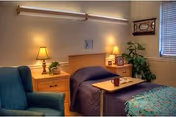 A cozy bedroom in a senior living facility featuring a single bed with a dark blue blanket, a wooden headboard, and a small overbed table with a cup on it. There are two wooden nightstands on either side of the bed, each with a lamp and decorative items. A comfortable blue armchair is positioned to the left, and a window with blinds is on the right wall. A framed picture and a green potted plant add to the room's homey atmosphere.