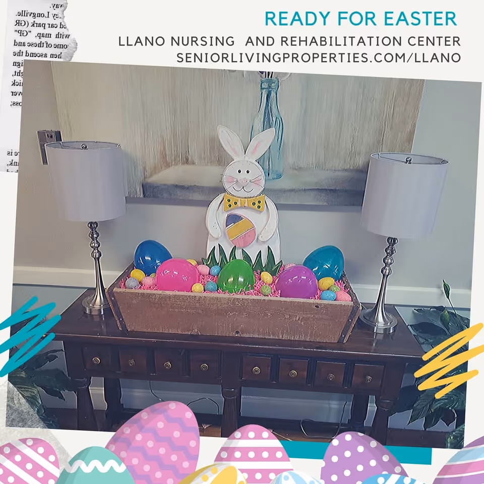 A decorated console table inside a nursing facility with an Easter bunny cutout, colorful plastic eggs, and two table lamps beneath a framed painting.
