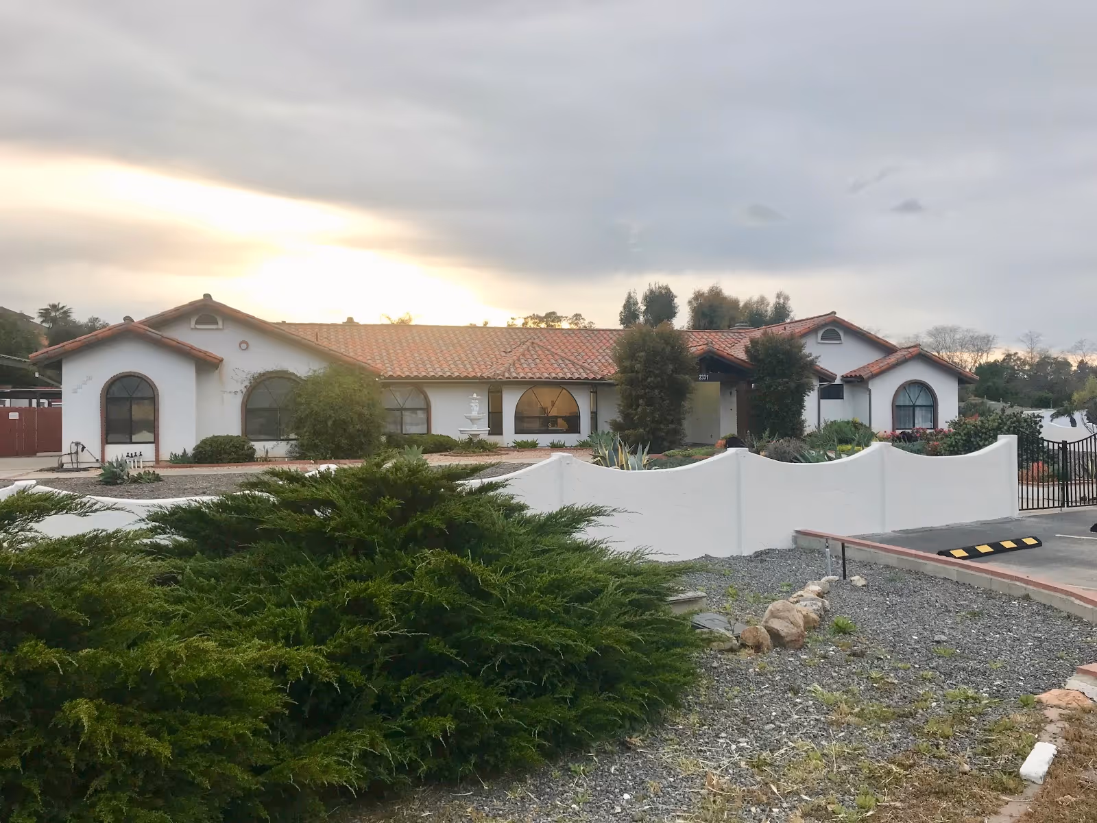 Exterior view of Monte Vista Manor, a single-story building with white walls and a red tile roof, surrounded by greenery and a white curved wall in front. The sky is cloudy with the sun setting in the background.