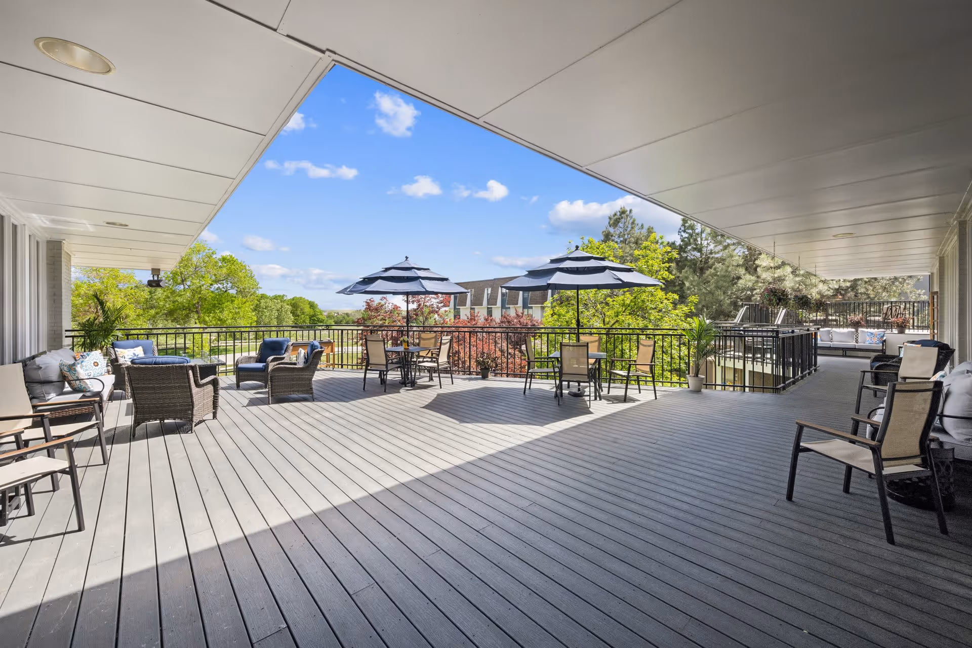 A spacious outdoor patio area with multiple seating arrangements including cushioned chairs, tables with umbrellas, and potted plants. The patio overlooks greenery and trees under a clear blue sky with some clouds.