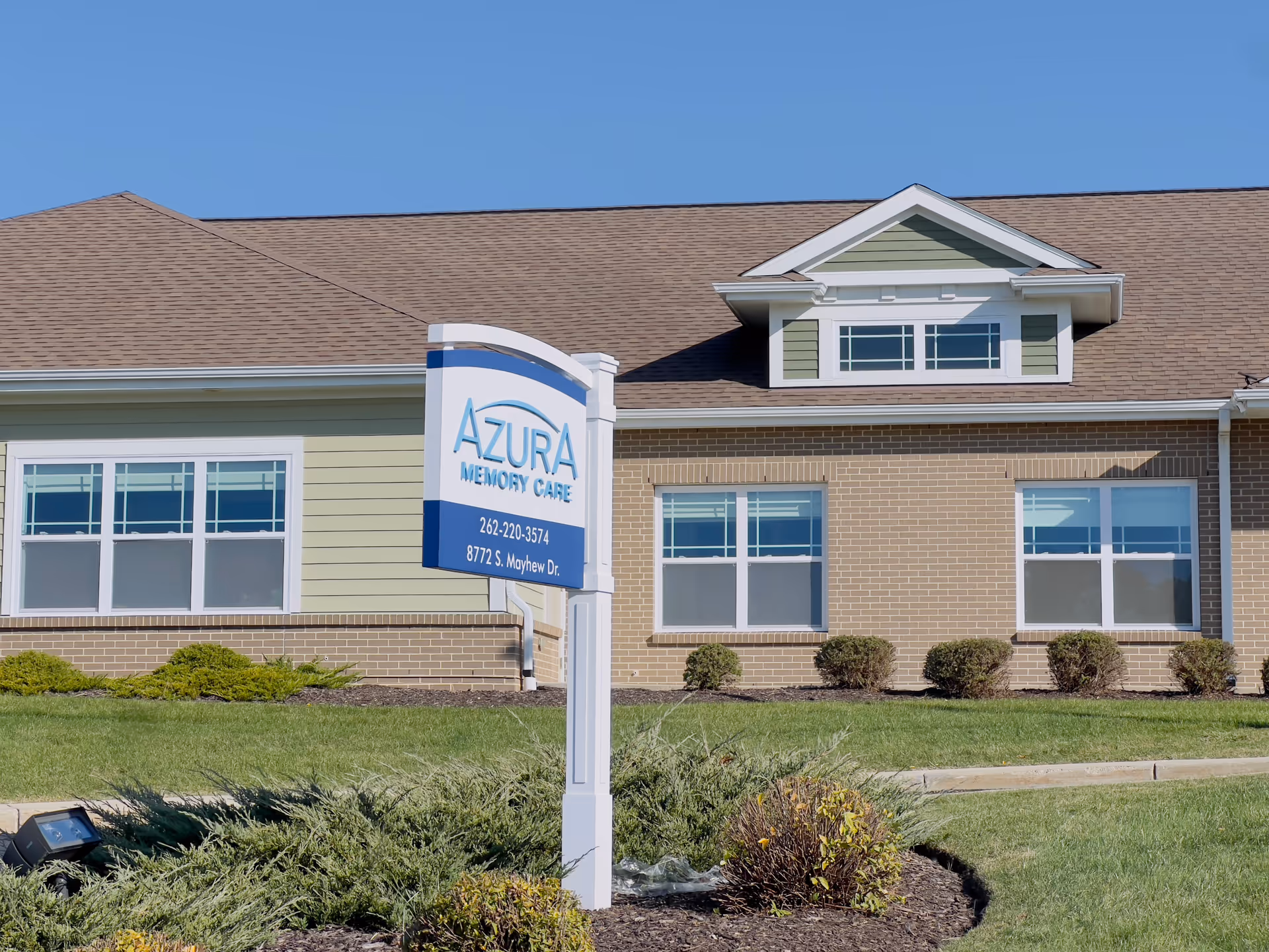 Exterior view of Azura Memory Care of Oak Creek building with a sign in front displaying the facility name, phone number, and address. The building has a brown shingled roof, light green and brick walls, and several windows. The surrounding area includes green grass, shrubs, and a clear blue sky.