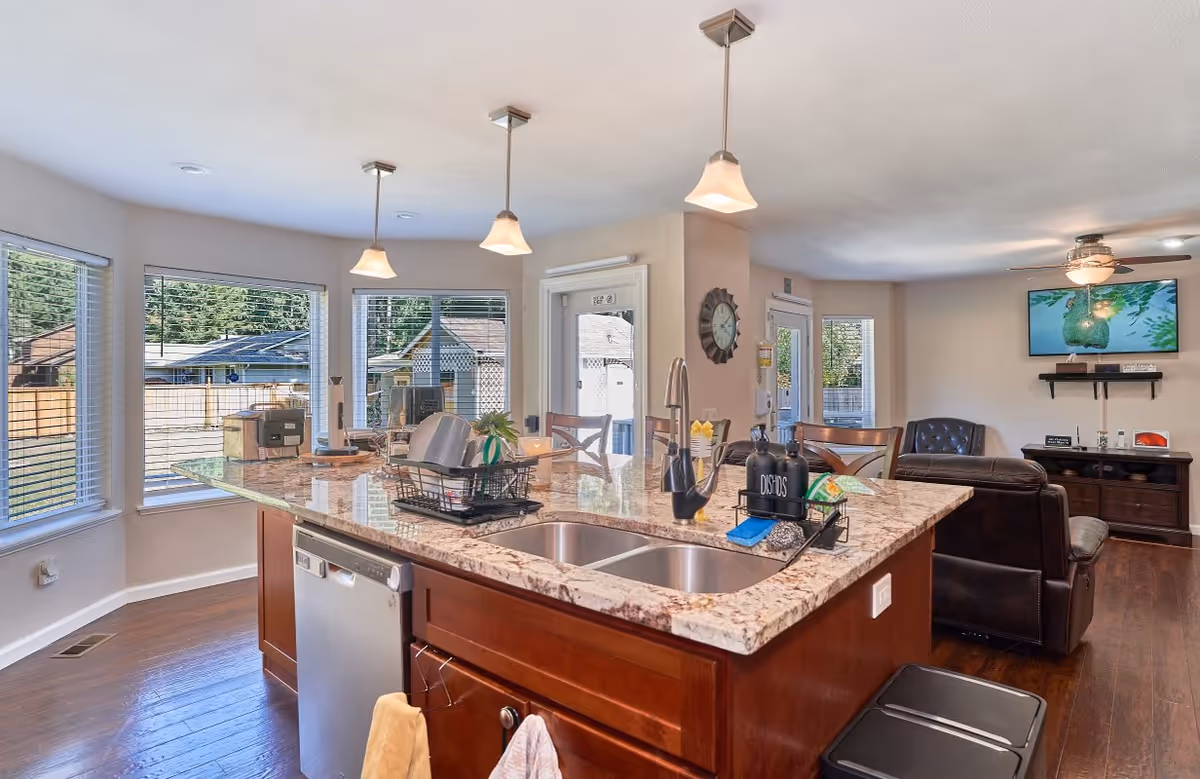 A bright and spacious kitchen area with a large marble countertop island featuring a double sink, dishwasher, and various kitchen items. The kitchen opens into a living room with a dark leather couch, a wall-mounted TV, and a ceiling fan. Large windows provide natural light and a view of the outdoors.