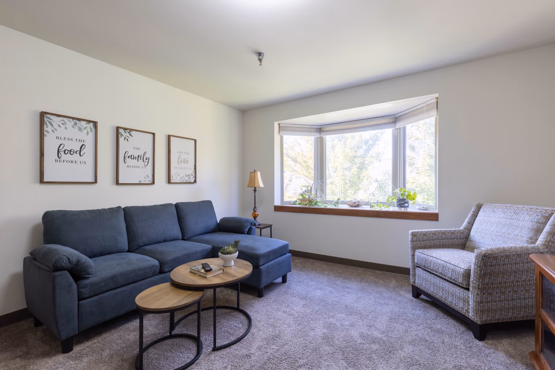 A cozy living room with a dark blue sectional sofa, a patterned armchair, and two nested round wooden coffee tables. There is a large bay window with plants on the windowsill letting in natural light. Three framed wall art pieces above the sofa read 'Bless the food before us', 'The family beside us', and 'And the love between us'. A floor lamp stands next to the sofa.
