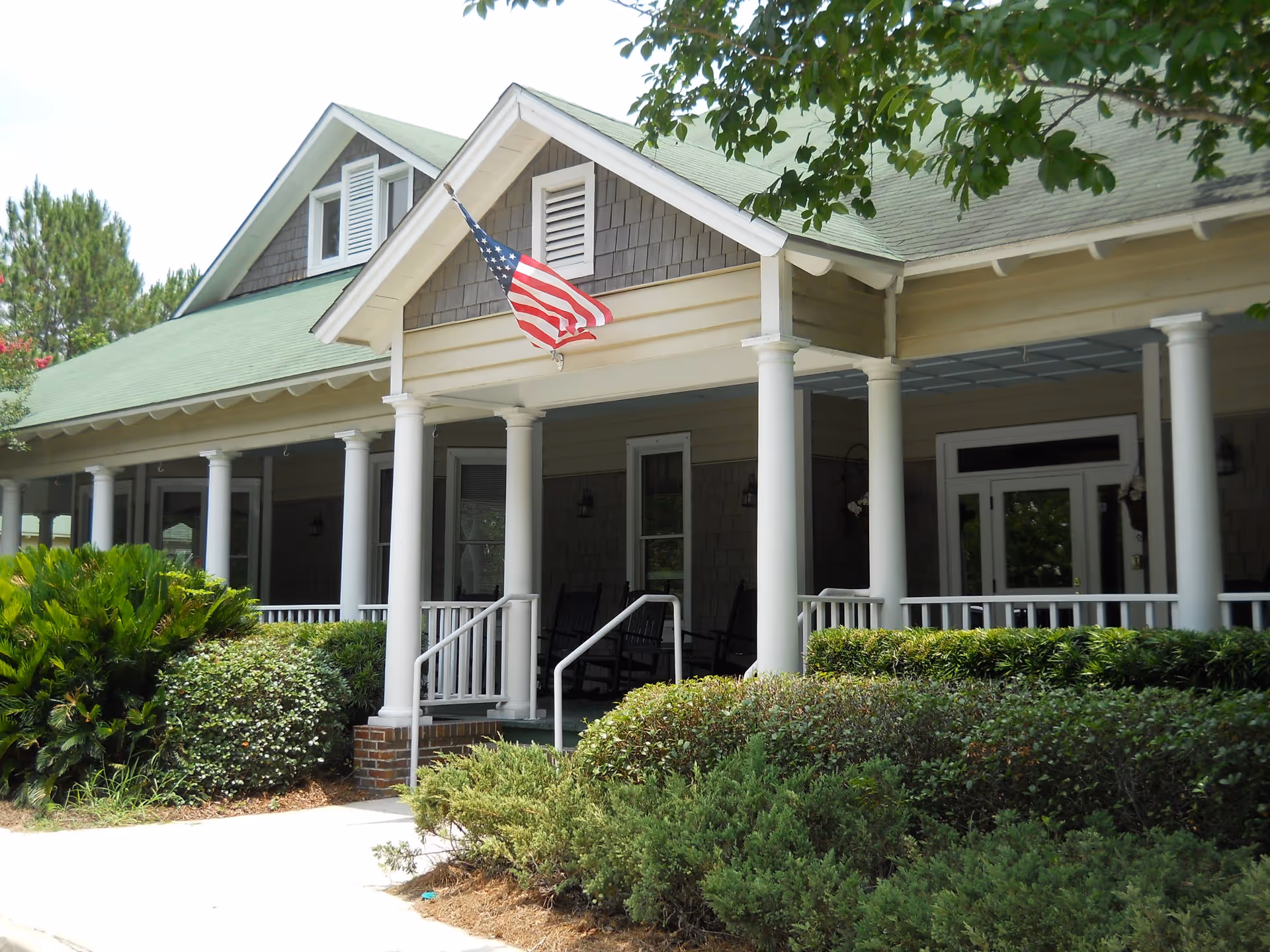 Front porch of a light-colored house with white columns, rocking chairs, an American flag, and landscaped shrubs.
