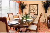 Dining room with a long wooden table set with plates and glasses, surrounded by upholstered chairs, potted plants, and wall decor.