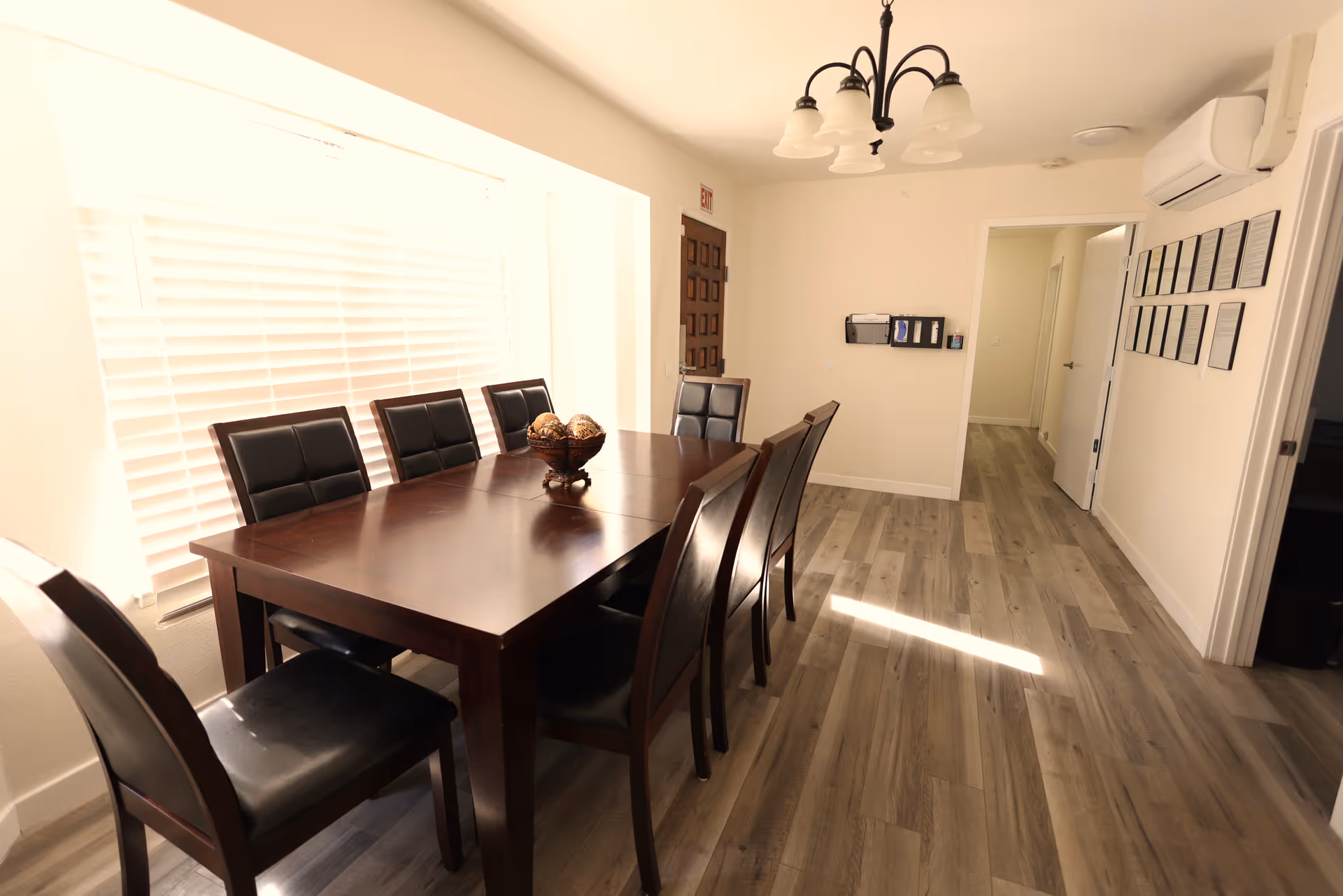 Sunlit dining room with a dark wooden table and eight leather-upholstered chairs, hardwood floors, and a chandelier.