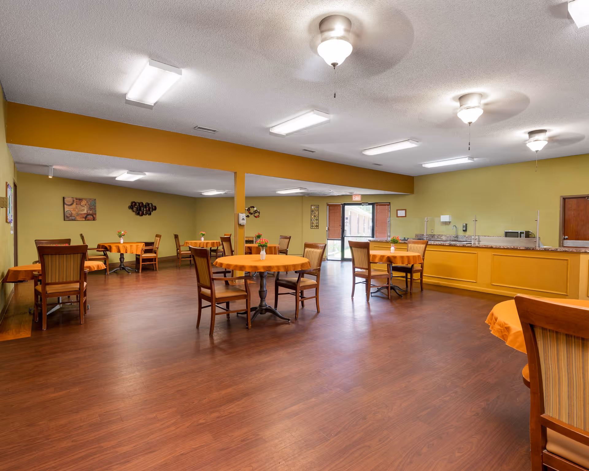 A spacious dining room with several round tables covered in orange tablecloths, each with a small flower vase centerpiece. Wooden chairs surround the tables. The room has wood flooring, yellow and green walls, ceiling fans with lights, and fluorescent ceiling lights. A counter area is visible on the right side, and a glass door leading outside is at the back.