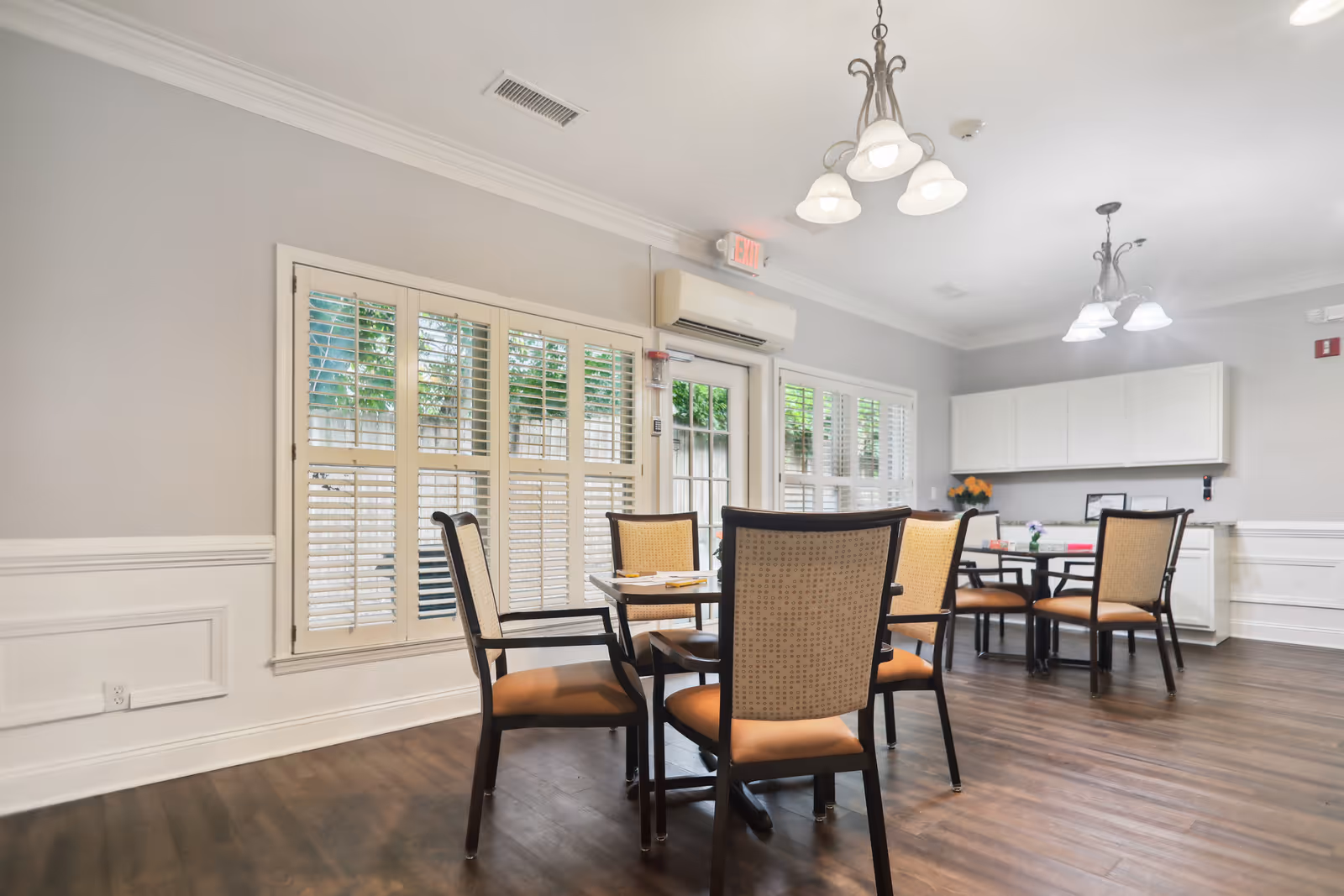 A bright dining room with wooden floors, several tables and chairs with cushioned seats, white walls with wainscoting, large windows with white shutters, and ceiling light fixtures.