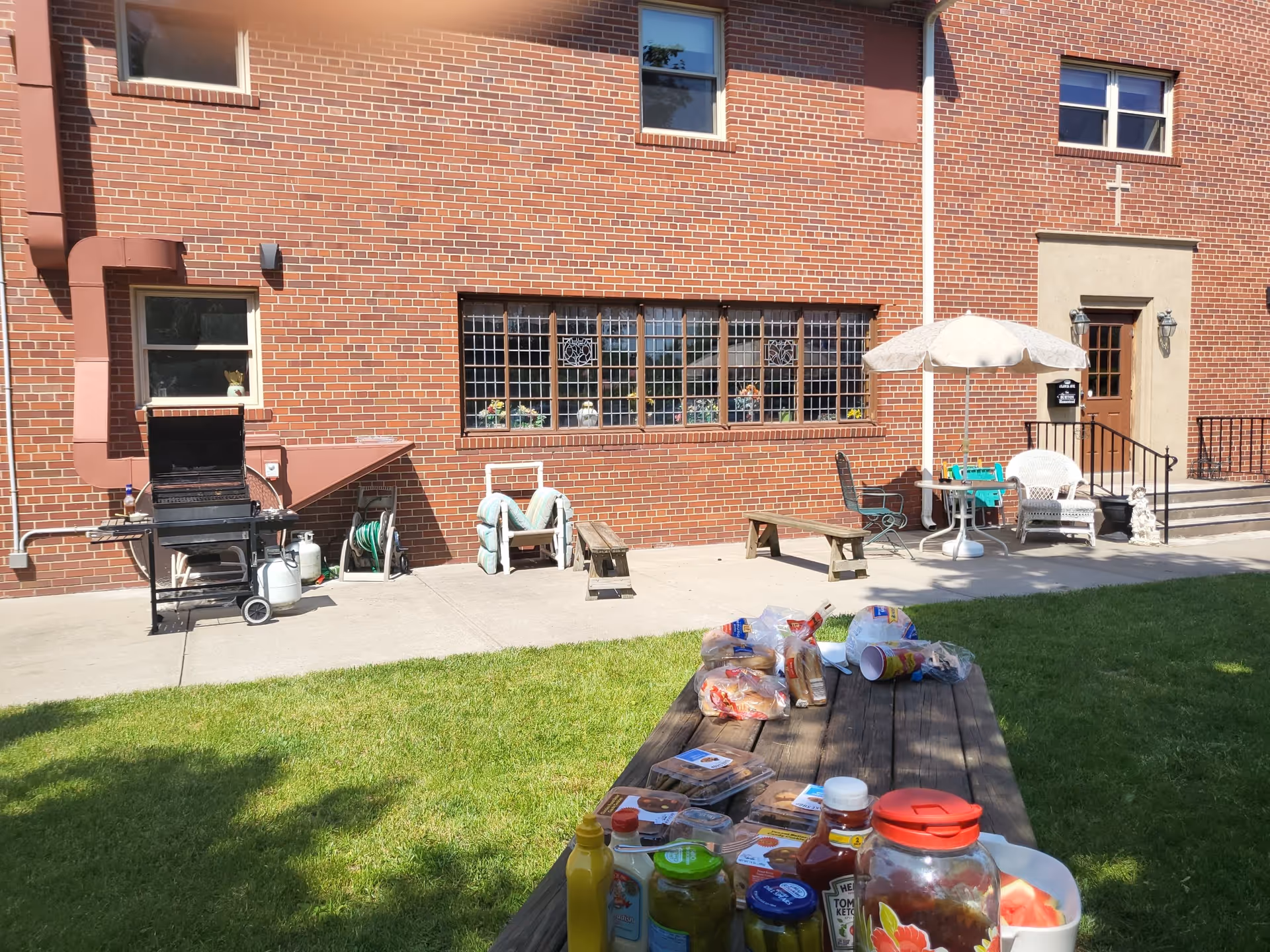 Outdoor patio area of a brick building with a barbecue grill, garden hose, benches, a table with an umbrella, and a picnic table with various condiments and food items on it, set on a grassy lawn.