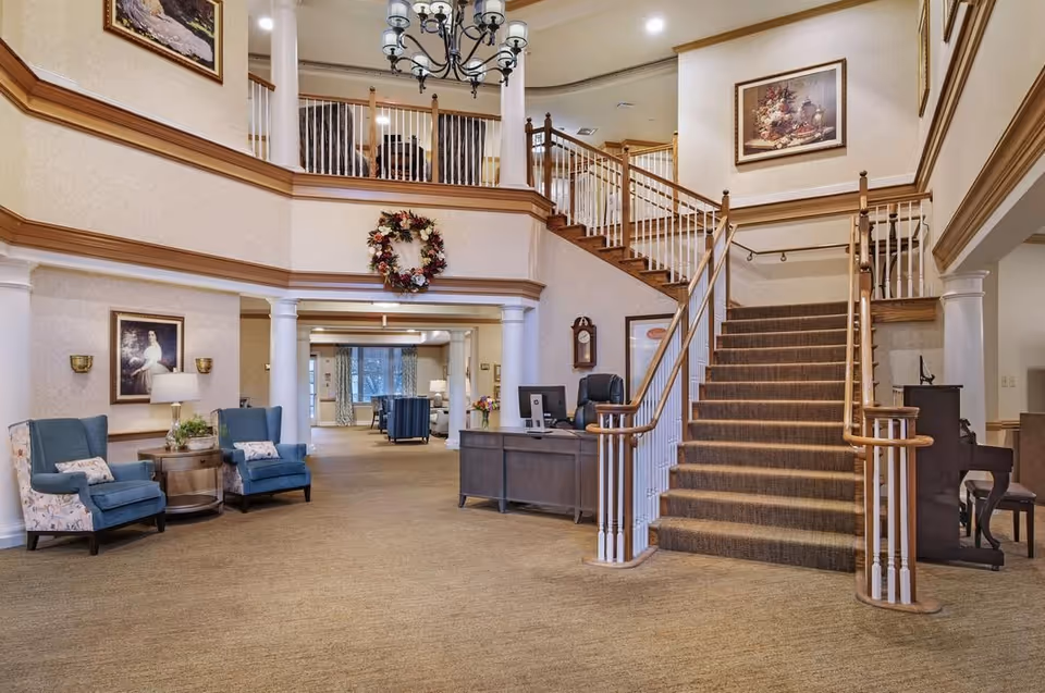 Spacious senior living lobby with a carpeted grand staircase, reception desk, and seating area under a chandelier.